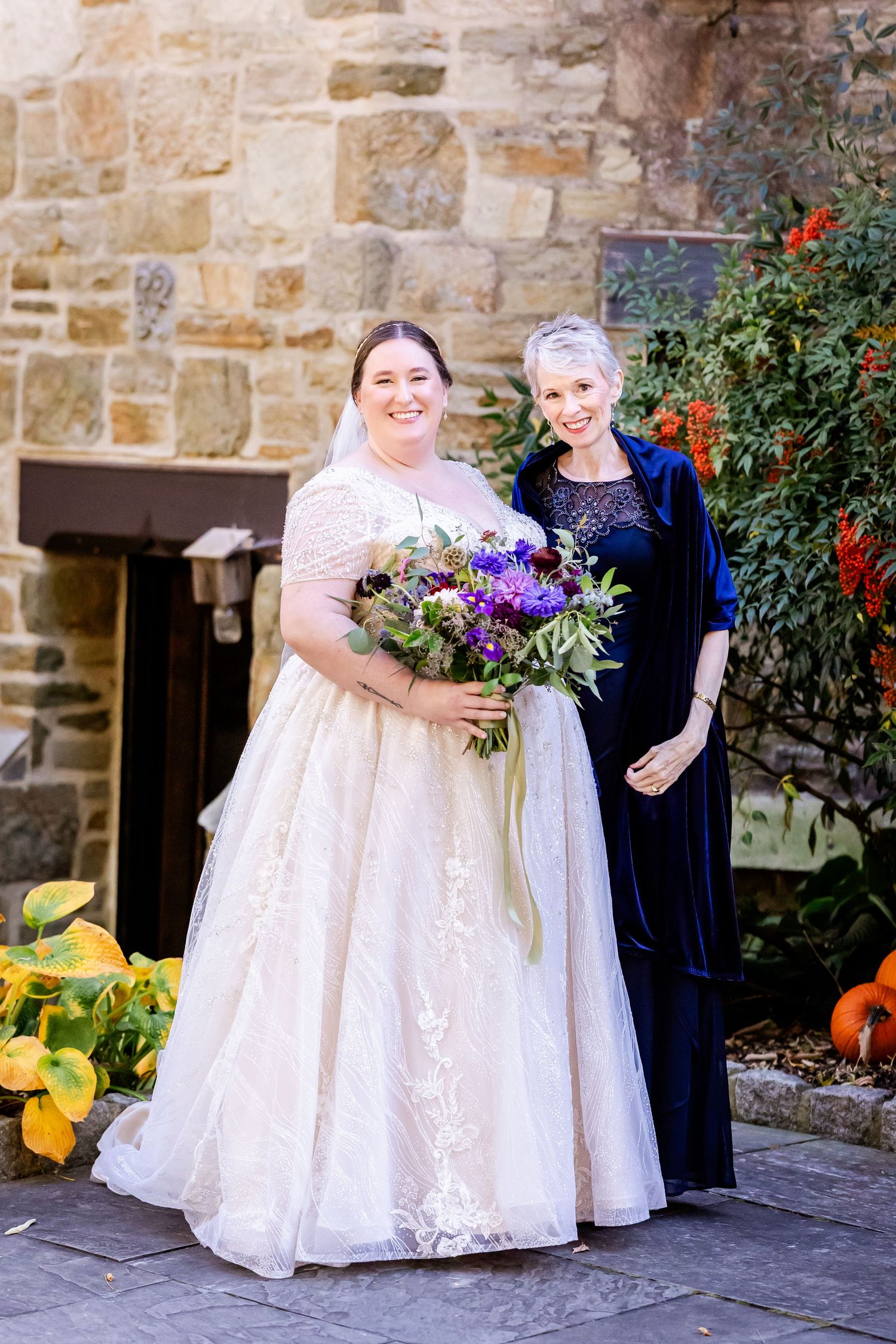 A pair stands together against stone wall backdrop with colorful autumn pumpkins and flowers.