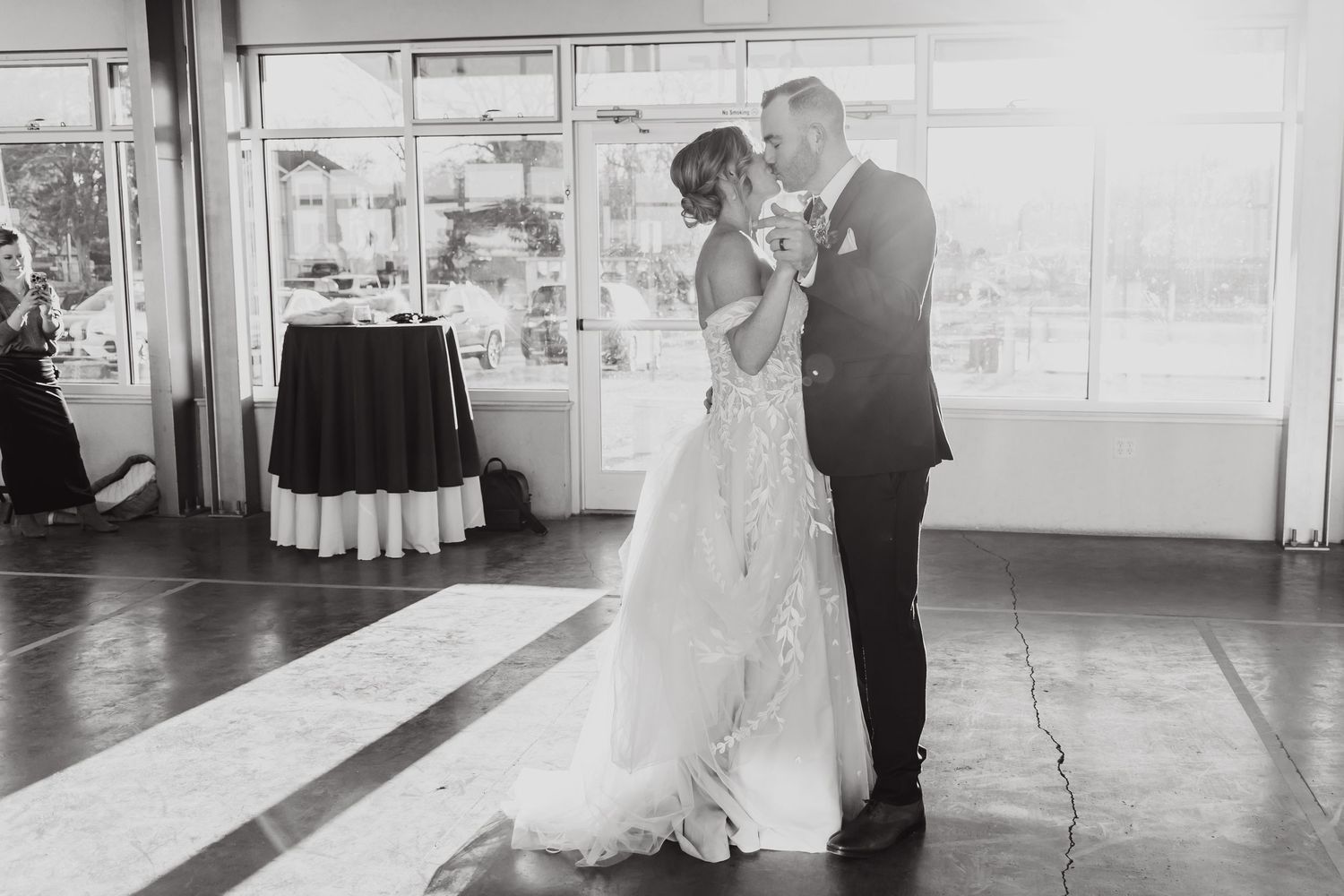 Black and white photo of couple dancing in elegant reception venue with large windows.