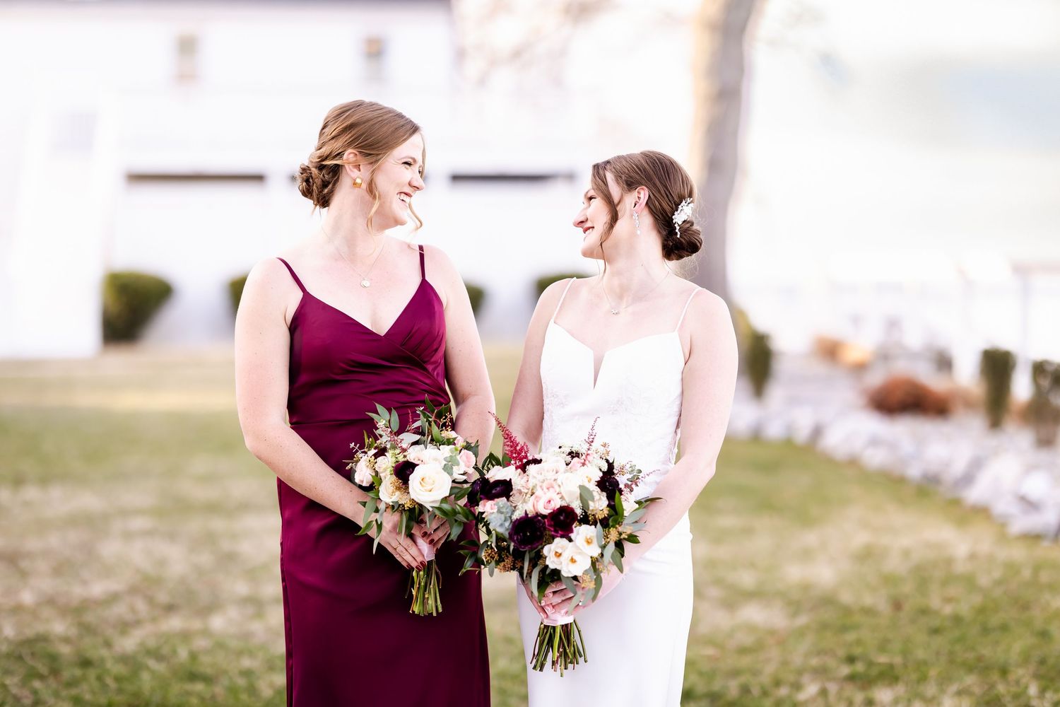 Two people in elegant dresses, one burgundy and one white, hold floral bouquets while standing together outdoors.