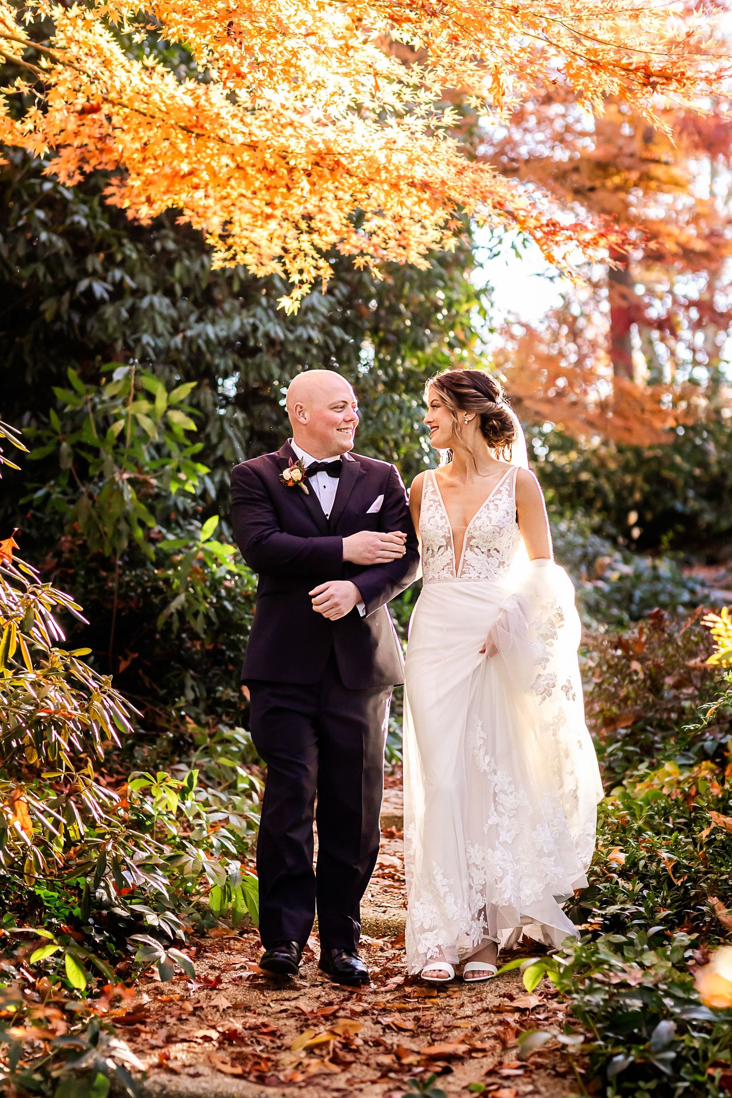 A couple walks arm in arm along a garden path surrounded by vibrant autumn foliage and falling leaves.