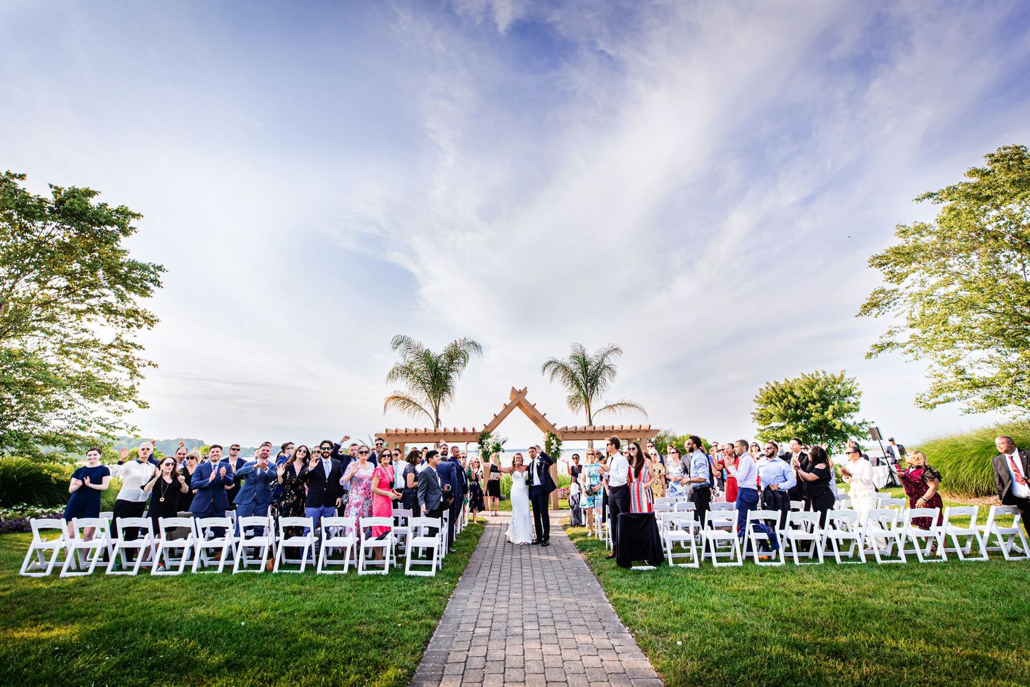 Outdoor garden wedding ceremony under dramatic sky with palm trees and white chairs lined up on green lawn.