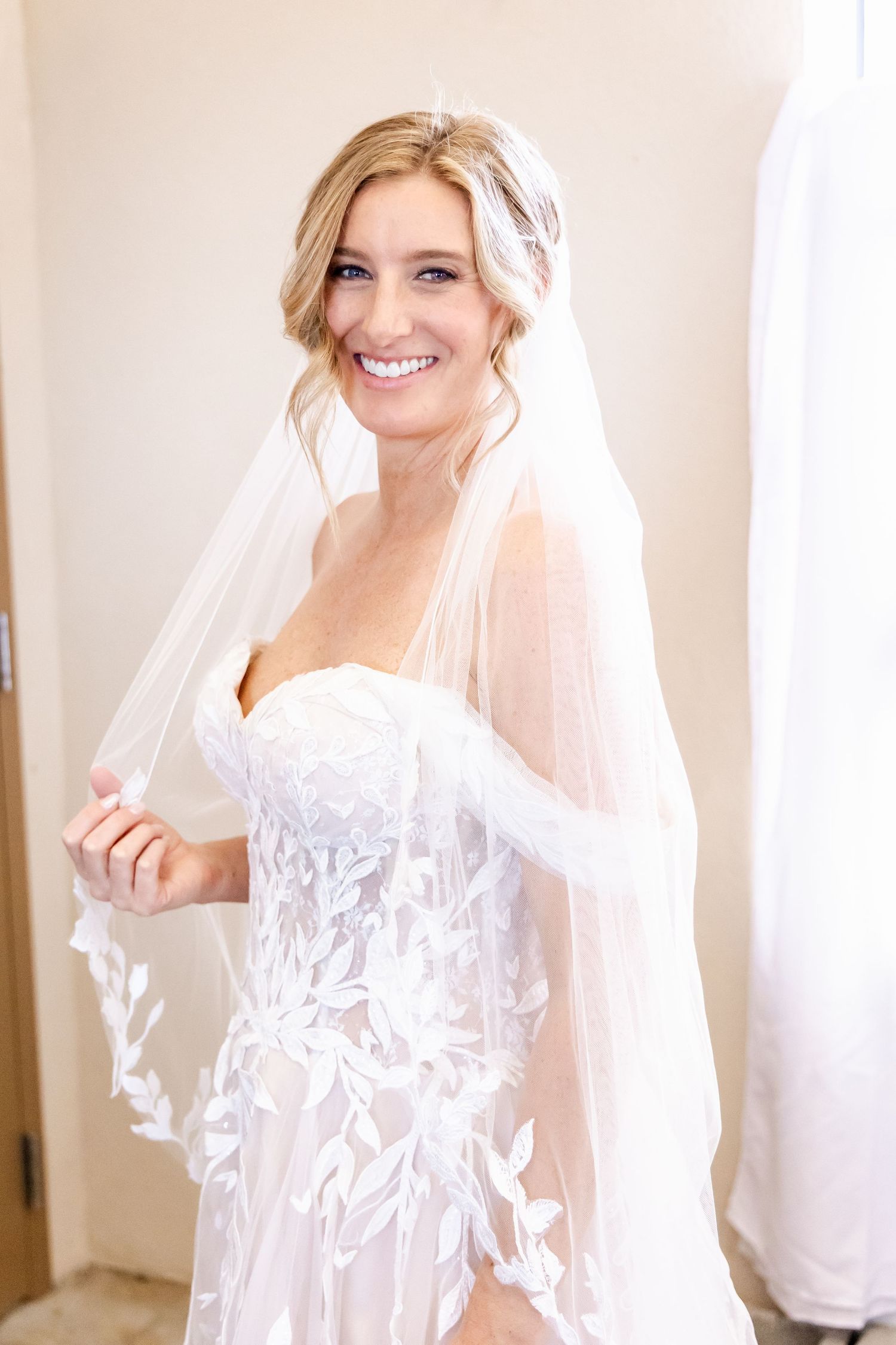 A bride wearing a beautiful white lace wedding dress with a flowing veil smiles in an indoor setting.