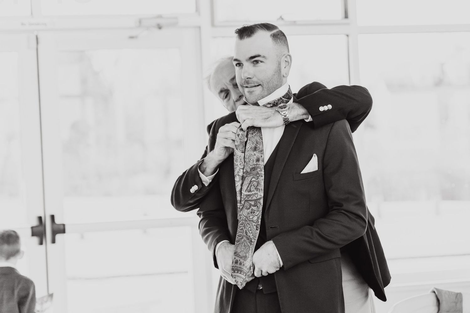 A formal black and white portrait of a groom adjusting his tie.