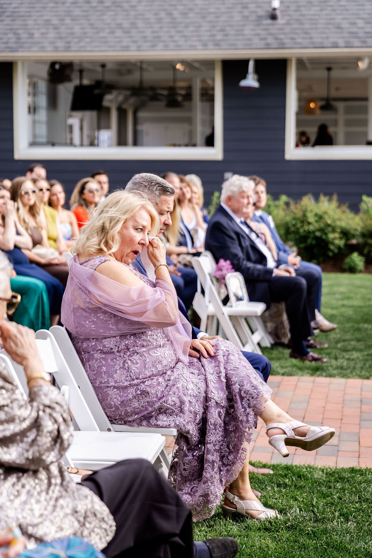 Wedding guests seated on white chairs watch outdoor ceremony on brick patio and lawn in afternoon light.
