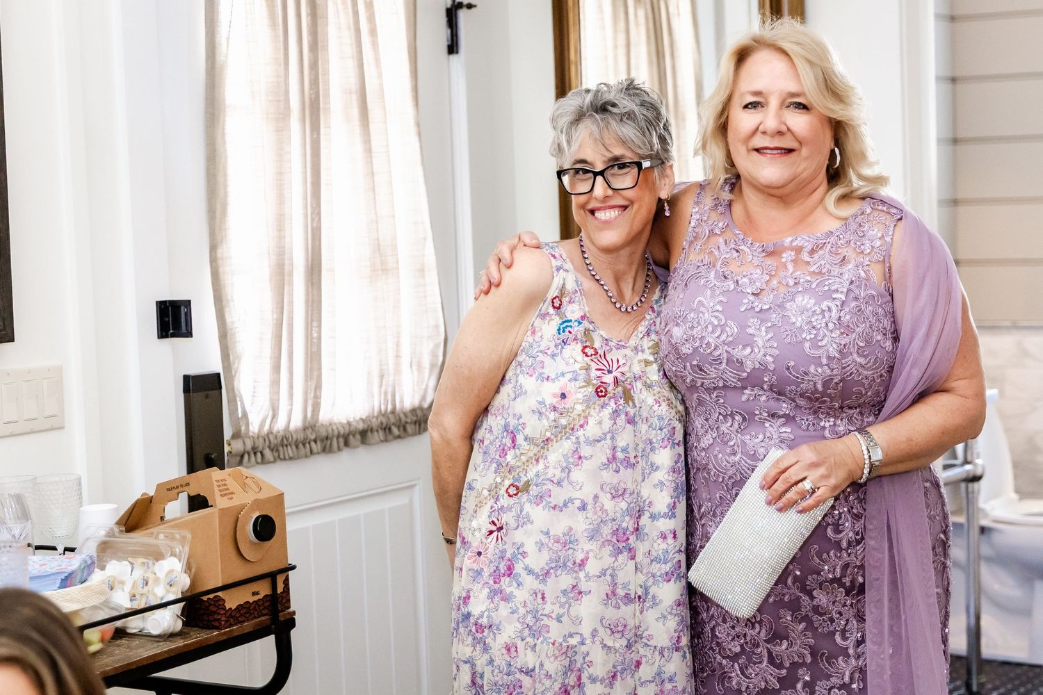 Two women in purple and floral dresses pose together during wedding celebration.