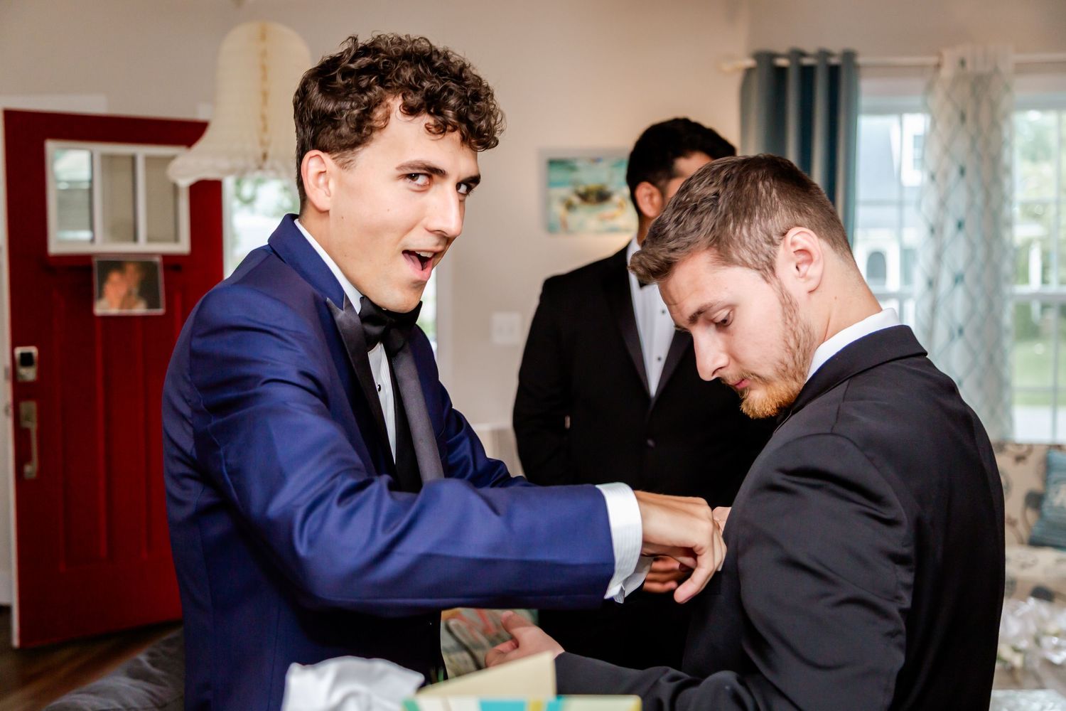Two men in suits helping each other with formal attire preparations in a room.