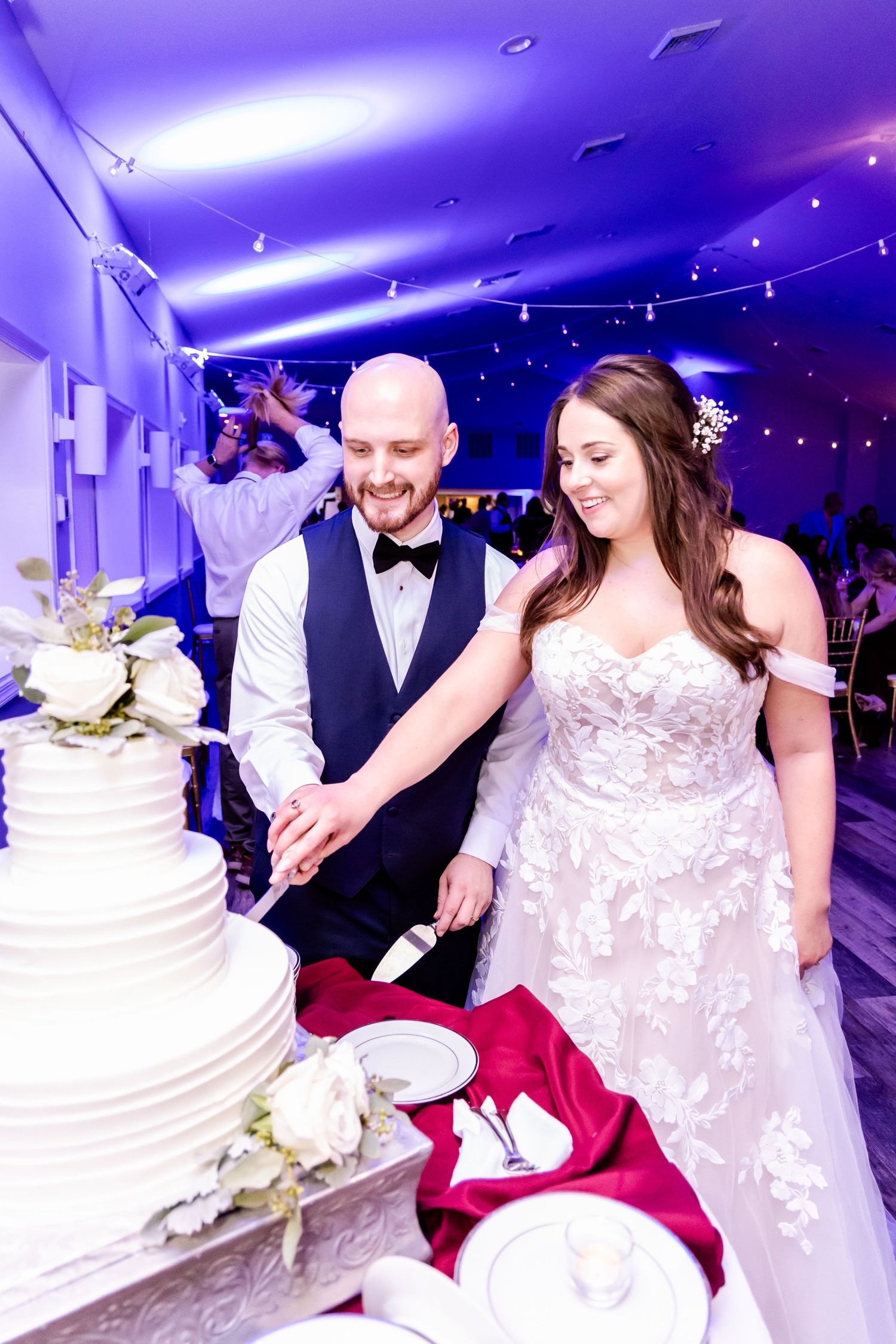 A couple cuts their elegant white wedding cake together under purple and blue lighting at their reception celebration.