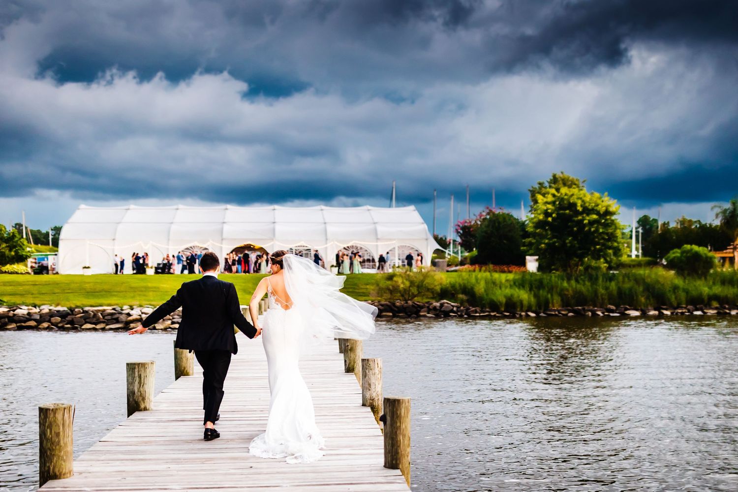 A couple in formal wedding attire walking together on a wooden dock over water with dramatic storm clouds overhead.