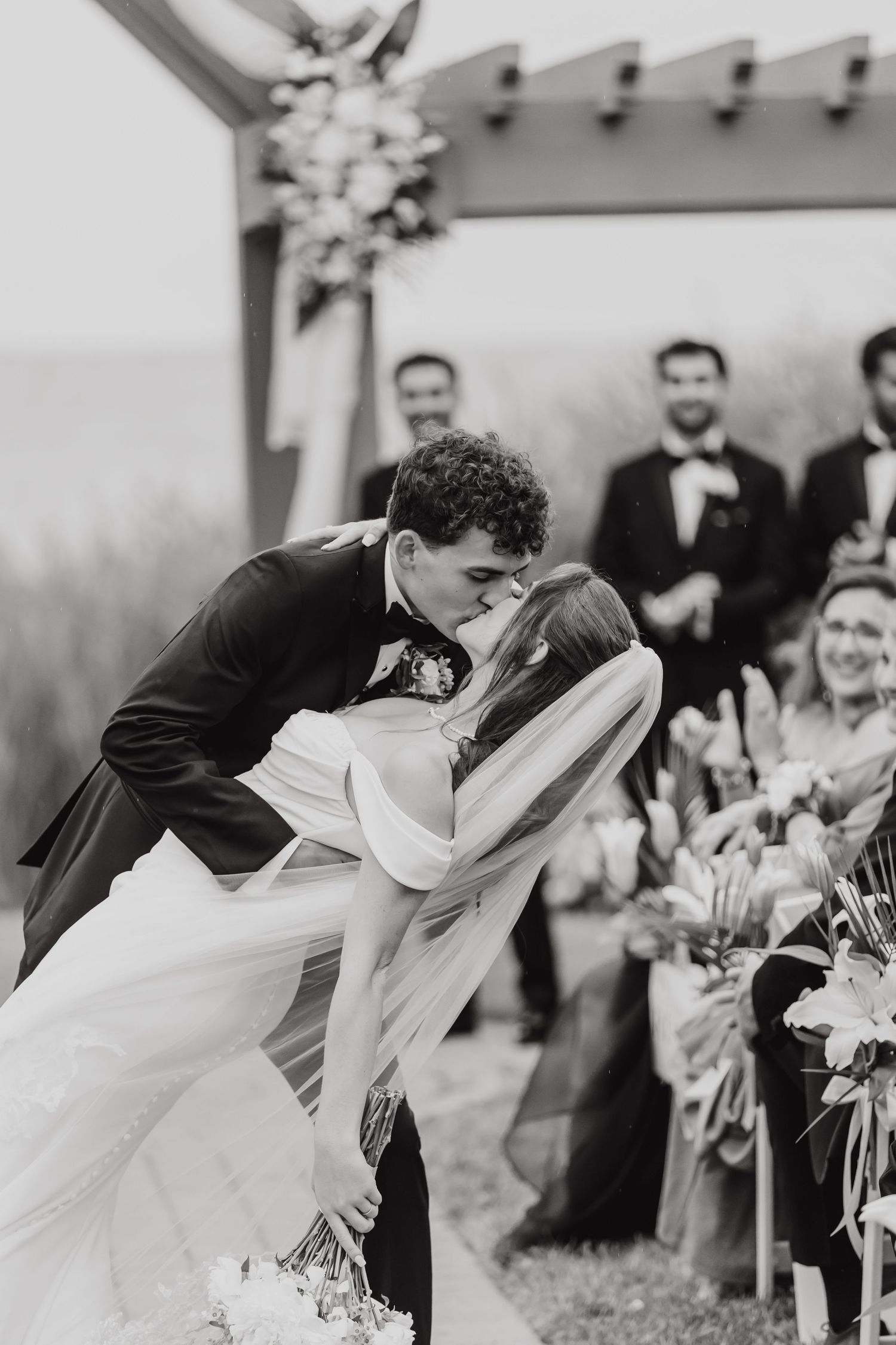 A romantic wedding ceremony kiss under a decorated outdoor pergola with guests looking on.