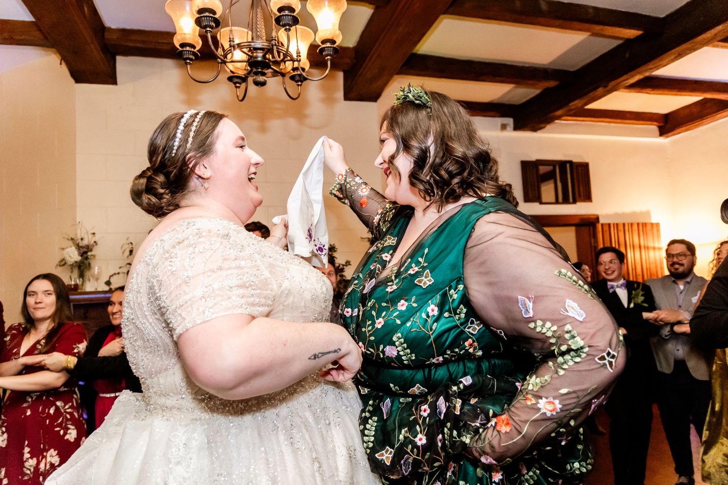 Two women in formal dresses share a joyful moment laughing together at an indoor celebration event.