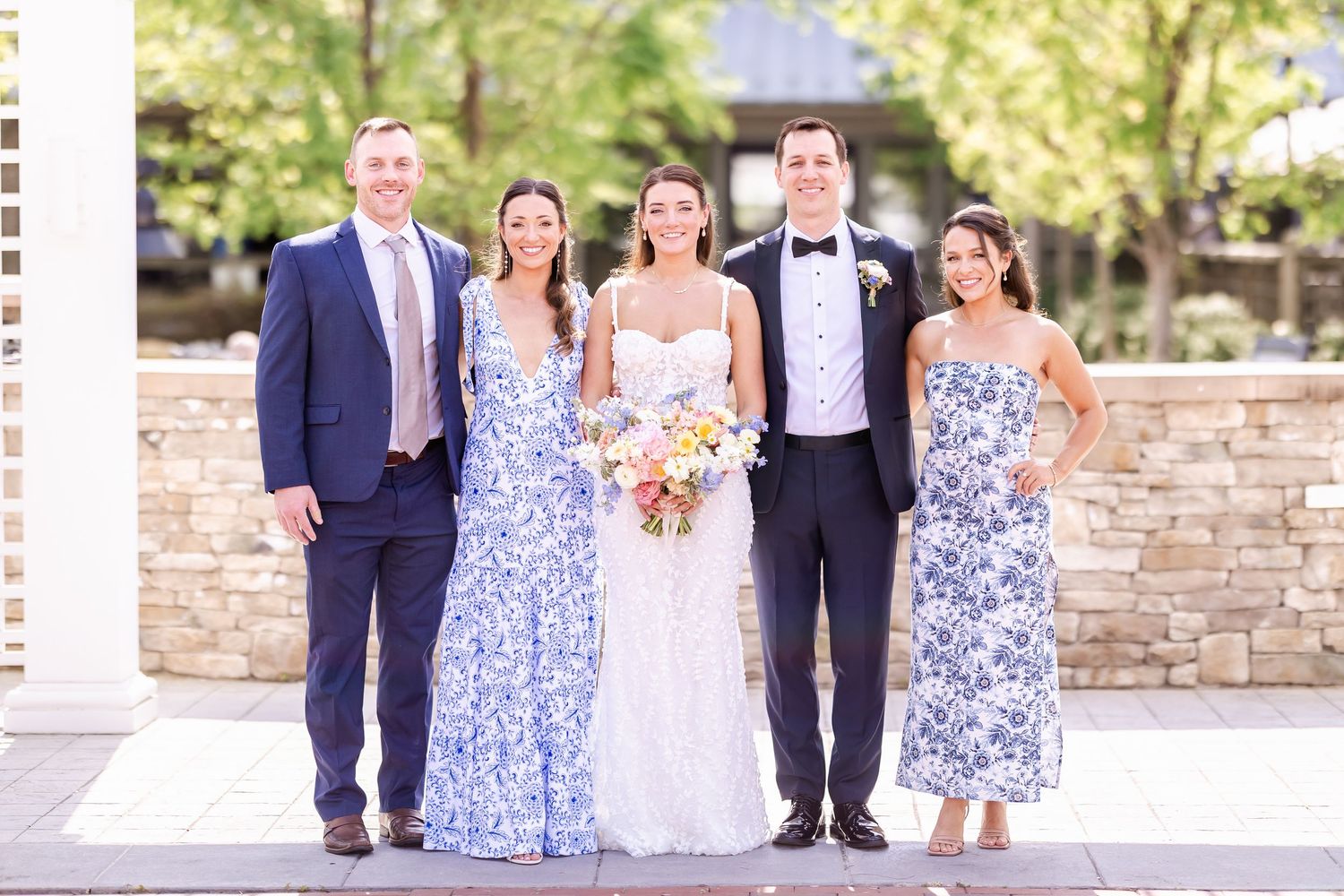 Wedding party members gathered outdoors near a stone wall.