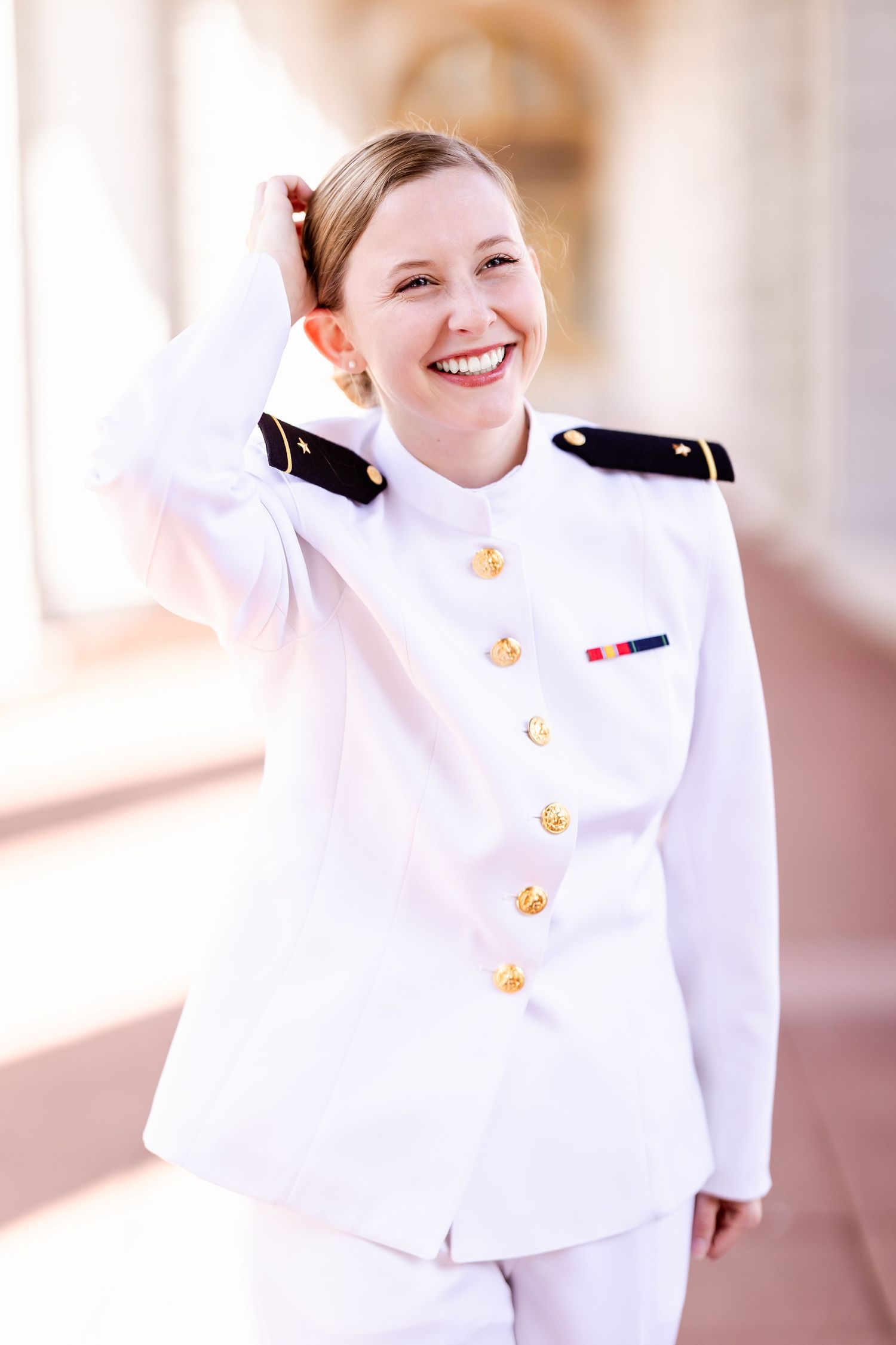 A service member in a crisp white dress uniform smiles warmly while standing in an elegant hallway.