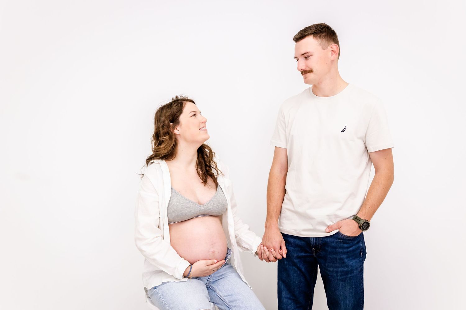 A pregnant woman in a white cardigan and gray top stands beside a person in a white t-shirt against a light background.