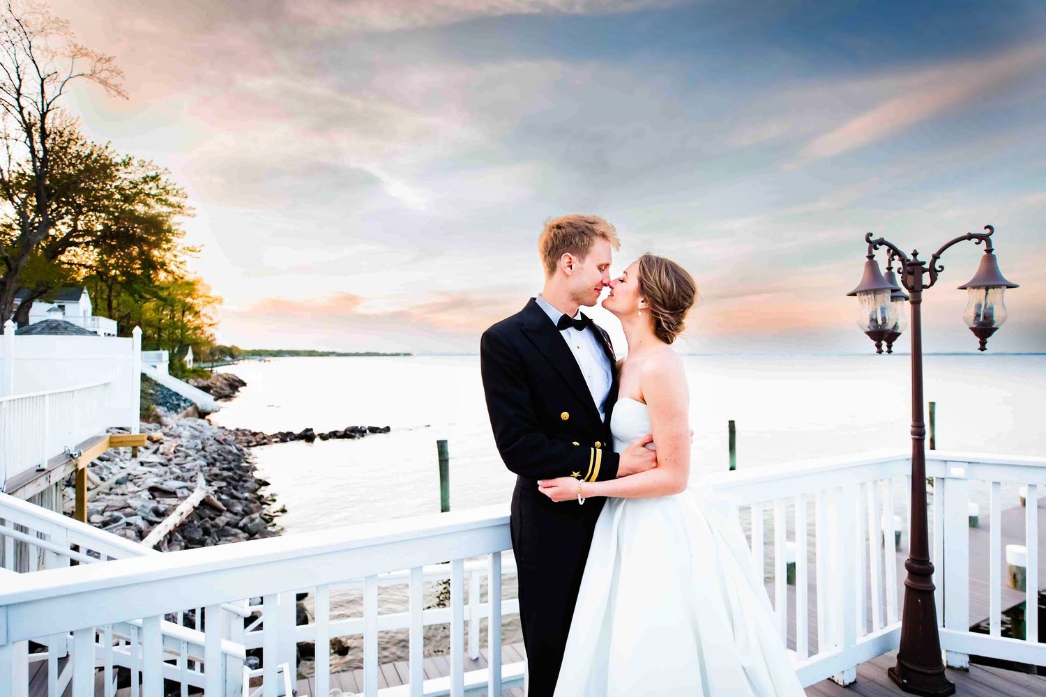 A couple shares a romantic moment on a white balcony overlooking a snowy winter beach at sunset with an ornate lamp post.