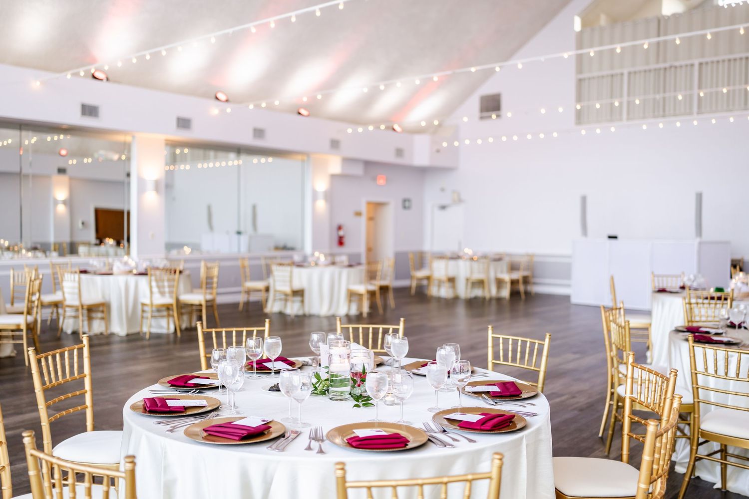 Elegant wedding reception venue with white and burgundy table settings and gold chiavari chairs.