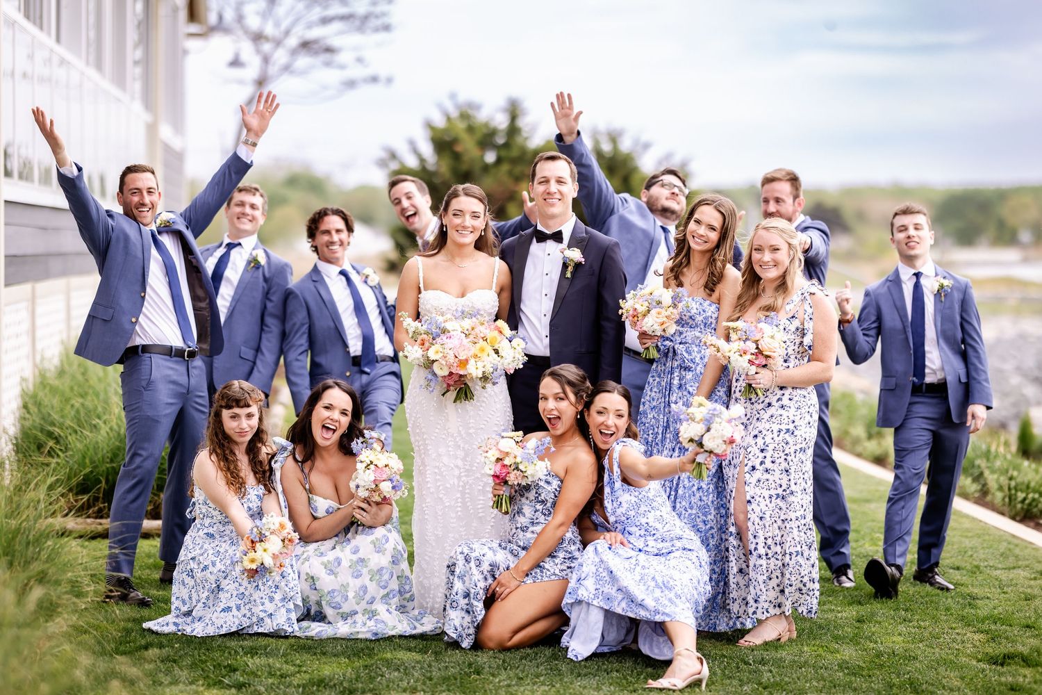 Wedding party in navy suits and floral blue dresses poses joyfully on a grassy lawn outside.
