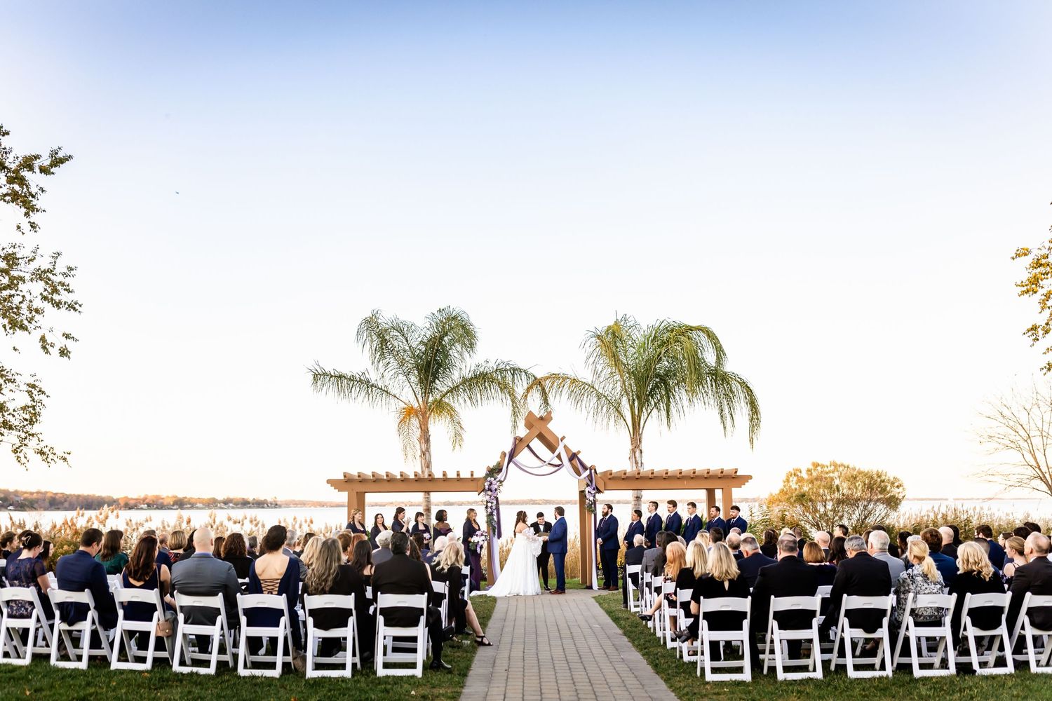 Outdoor waterfront wedding ceremony setup with white chairs and palm trees in background.