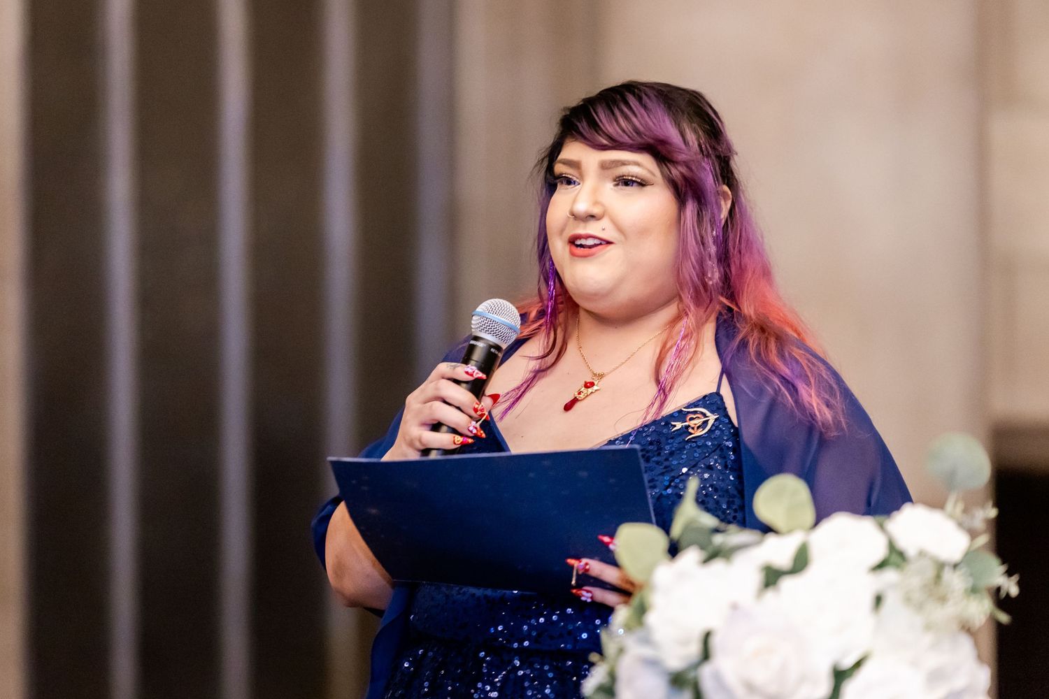 Guest in navy sequin dress gives speech at wedding reception with floral decorations.