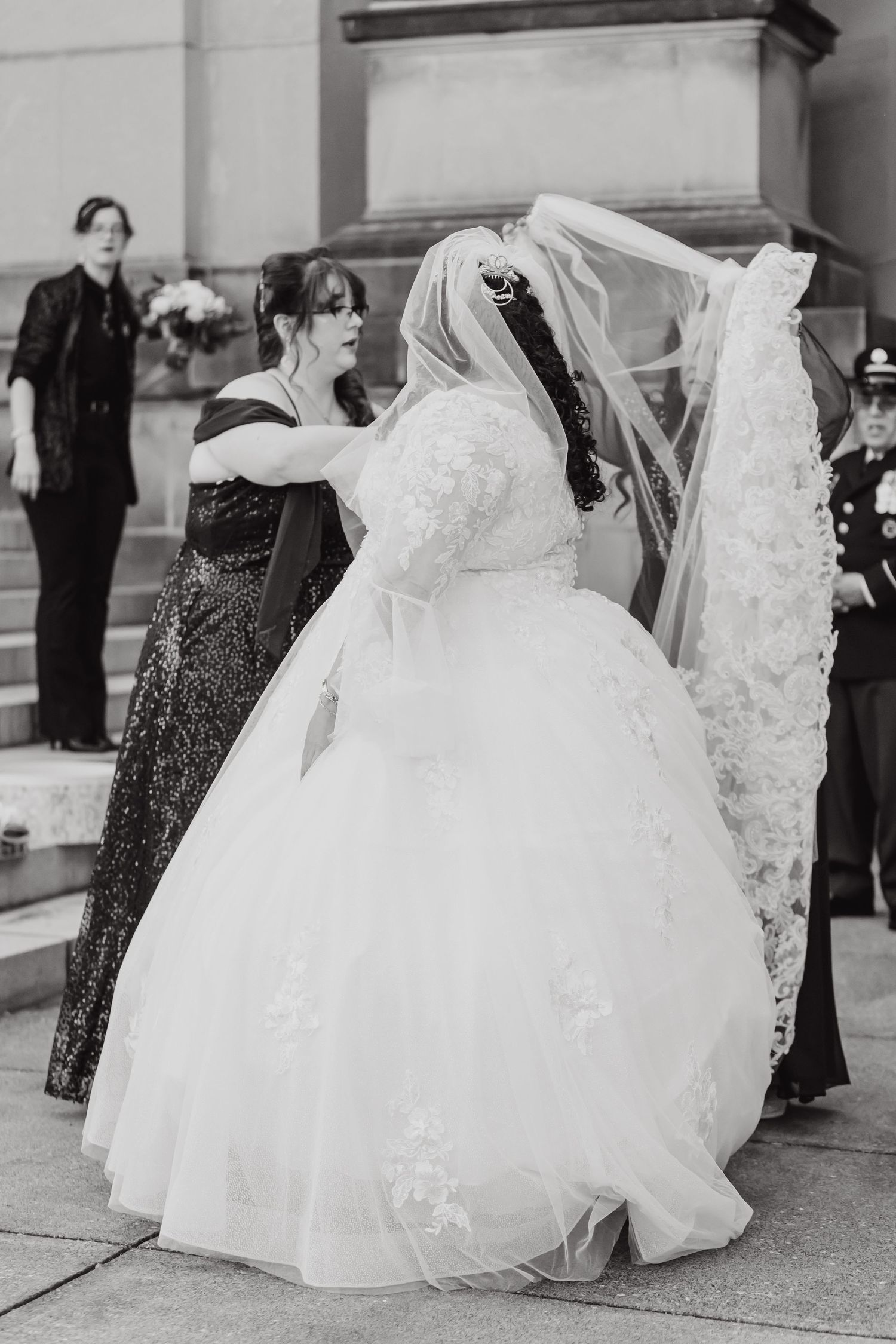 Black and white photo of bride in full ballgown being assisted with her dress on stone steps.