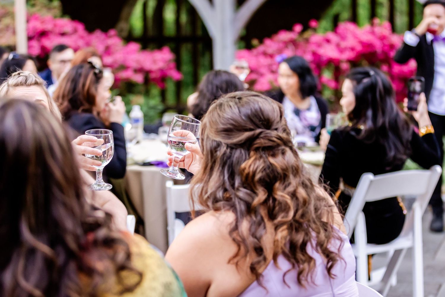 A speaker entertains wedding guests during reception toasts outdoors surrounded by pink flowering bushes.