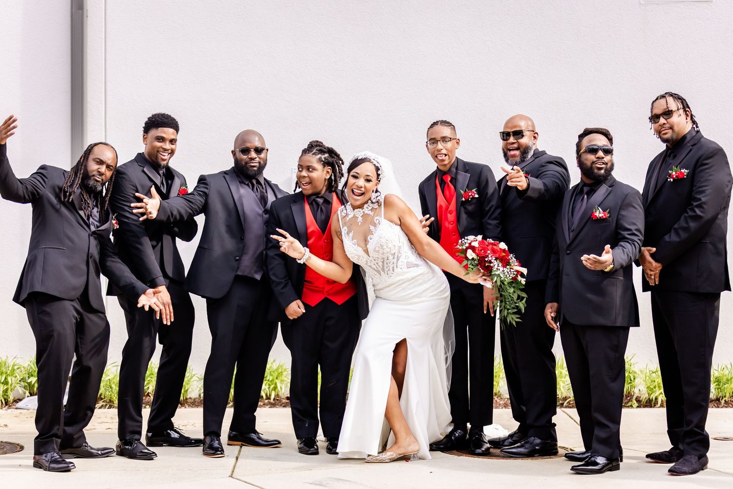 Groomsmen in black suits pose with newlyweds against white backdrop.
