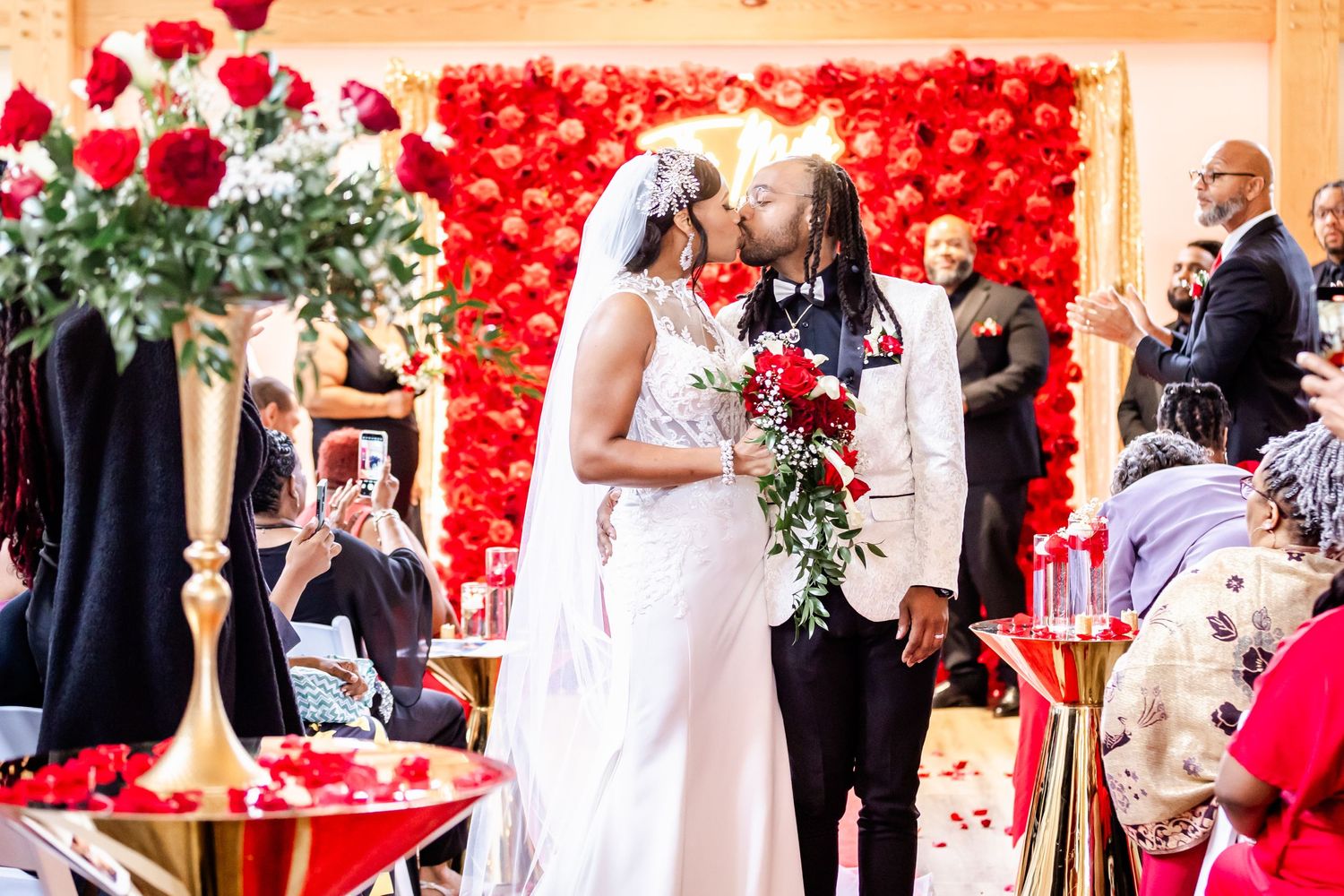 A wedding ceremony in front of a vibrant red rose backdrop with guests looking on.