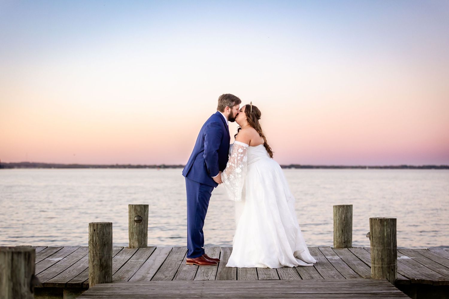 A romantic sunset wedding portrait of a couple on a wooden pier over water.