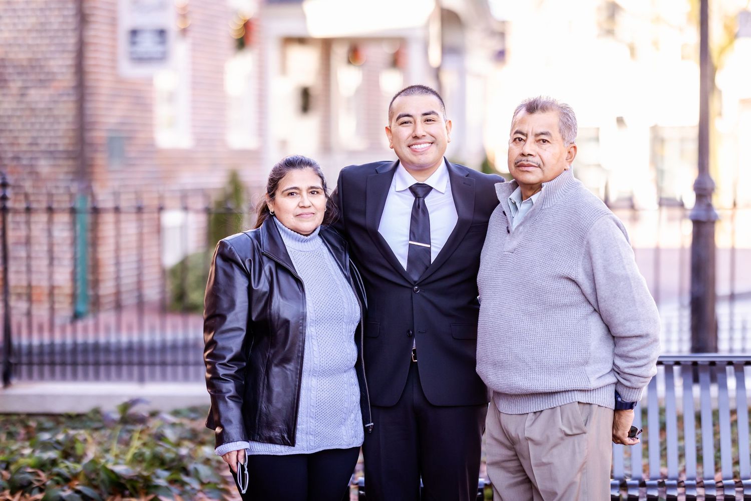 Three people pose together in front of iron fence and brick building.