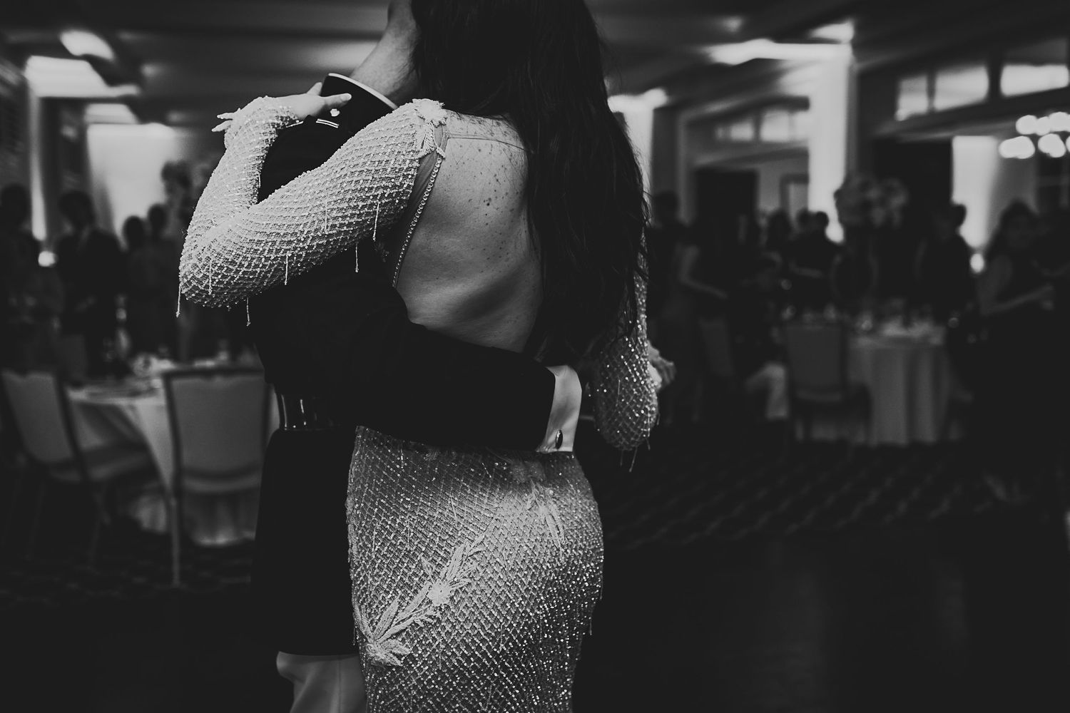 A couple embraces on a dimly lit dance floor, with the person wearing a sparkly sequined dress in black and white.