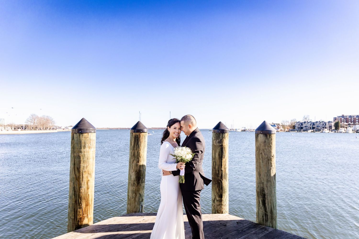 Newlyweds share a tender moment on a dock surrounded by wooden posts and waterfront views.