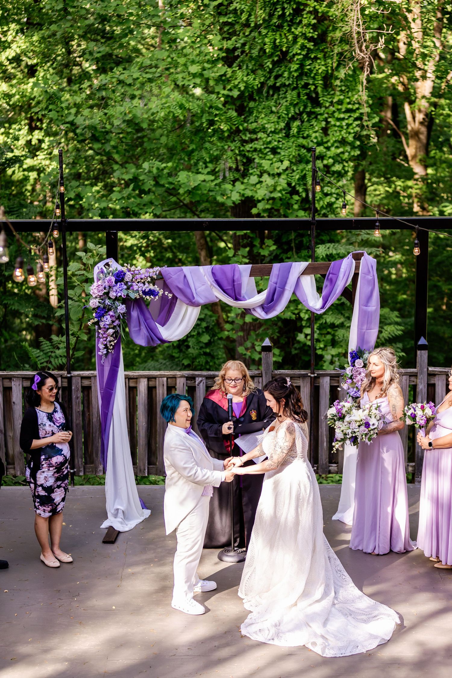 A wedding ceremony takes place under a purple-draped arch with floral decorations in a forest setting.