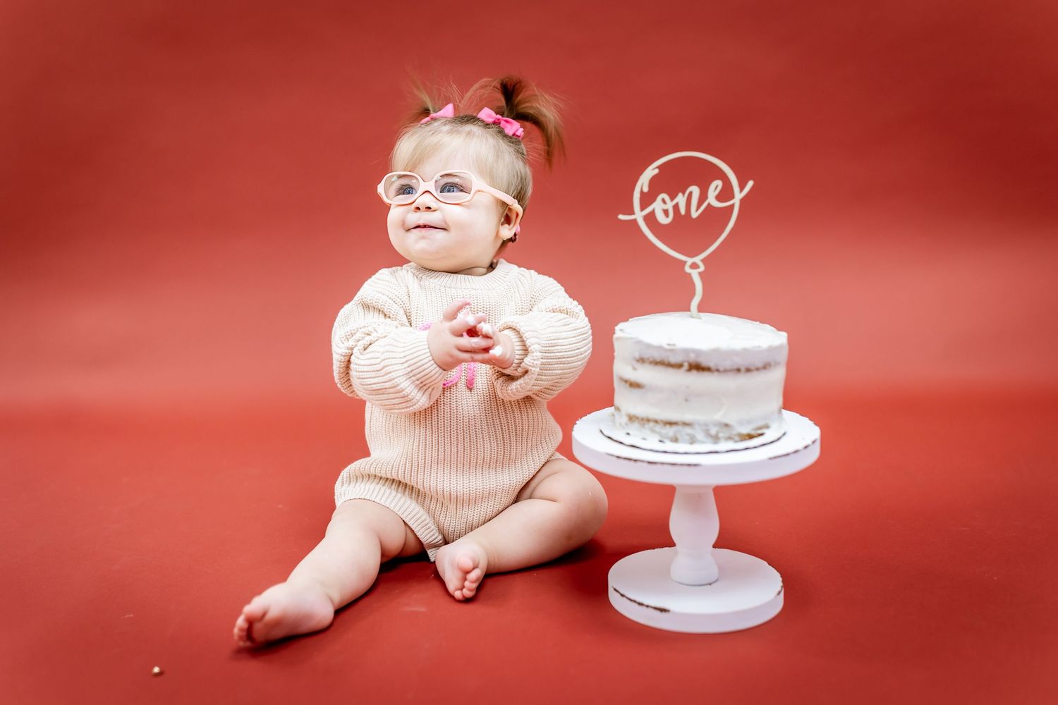 Baby in cream outfit sits near decorated white cake on red background.
