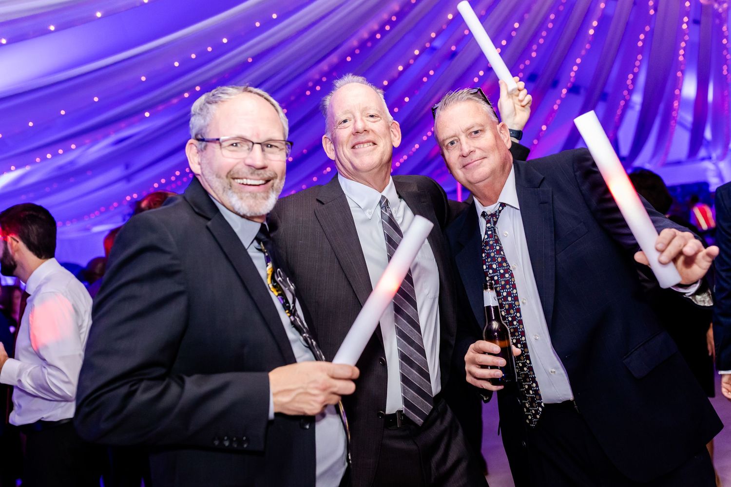 Wedding guests celebrating with glow sticks under purple draped ceiling.