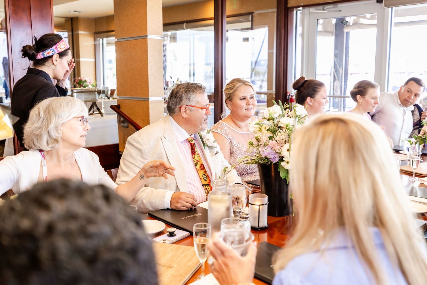 Wedding reception guests laugh and celebrate at a restaurant table decorated with floral centerpieces.