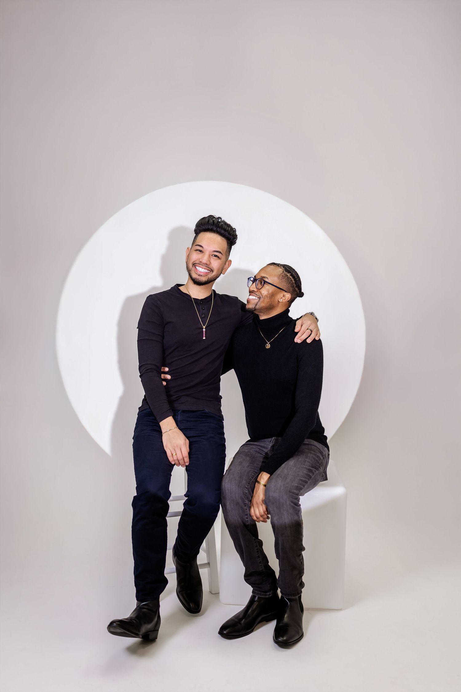 Two friends in dark clothing pose playfully while sitting on a curved white bench during a photo session.