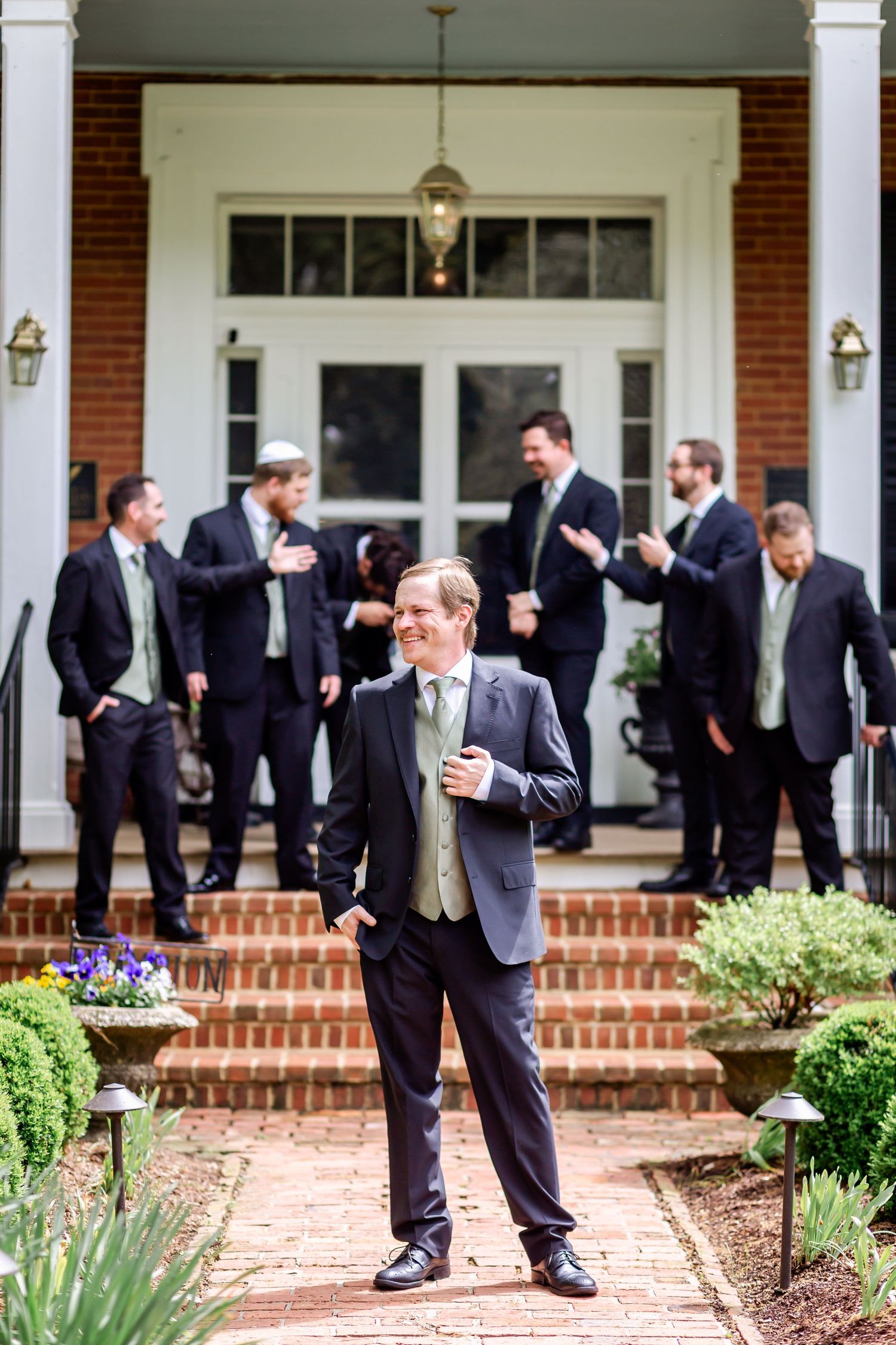 Wedding guests in dark suits gather on a brick porch with white columns and landscaping at an elegant venue.
