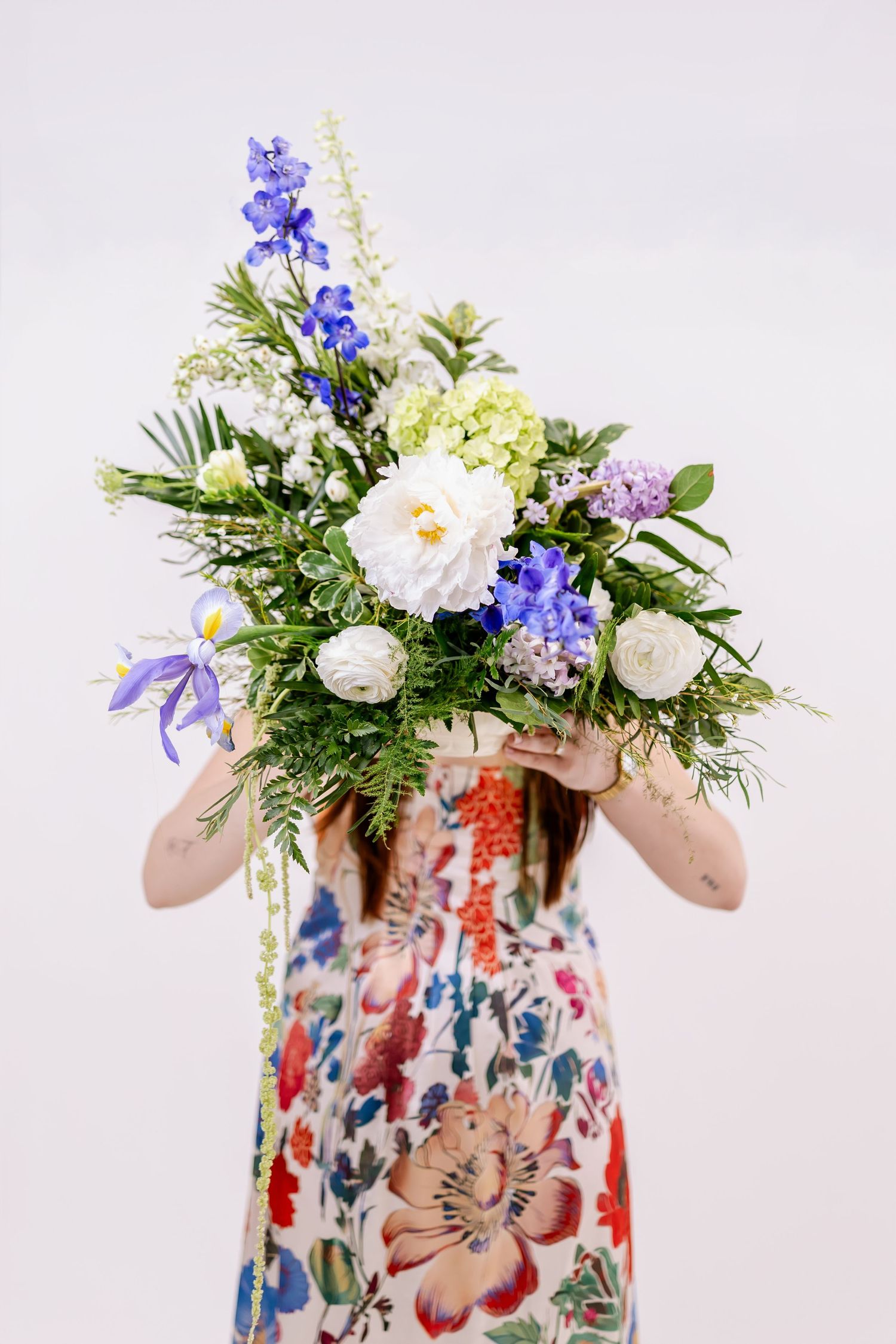 Woman in floral dress holding a large bouquet of blue, white, and green flowers covering her face.