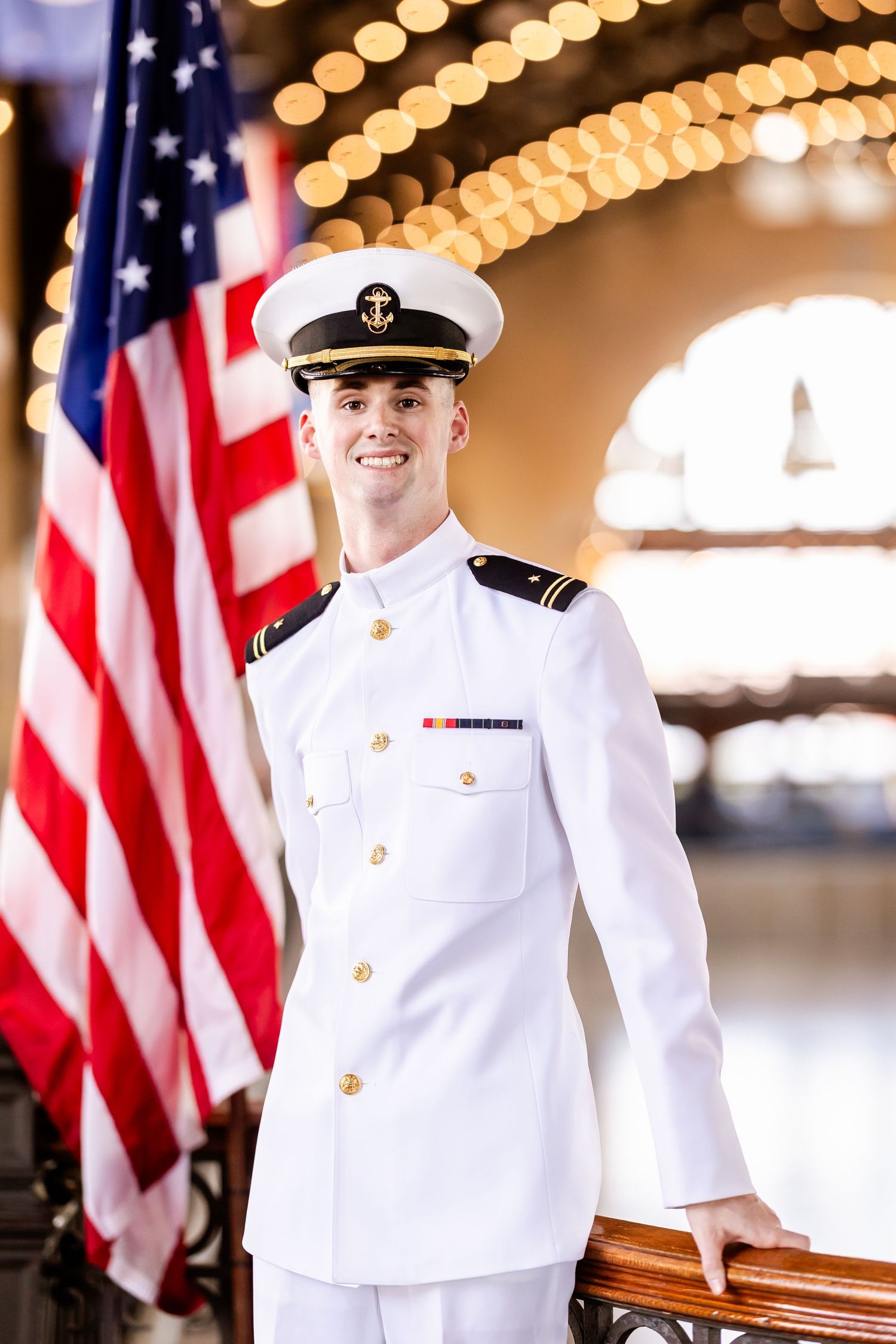 Young naval officer in white dress uniform smiling beside American flag in elegant venue.