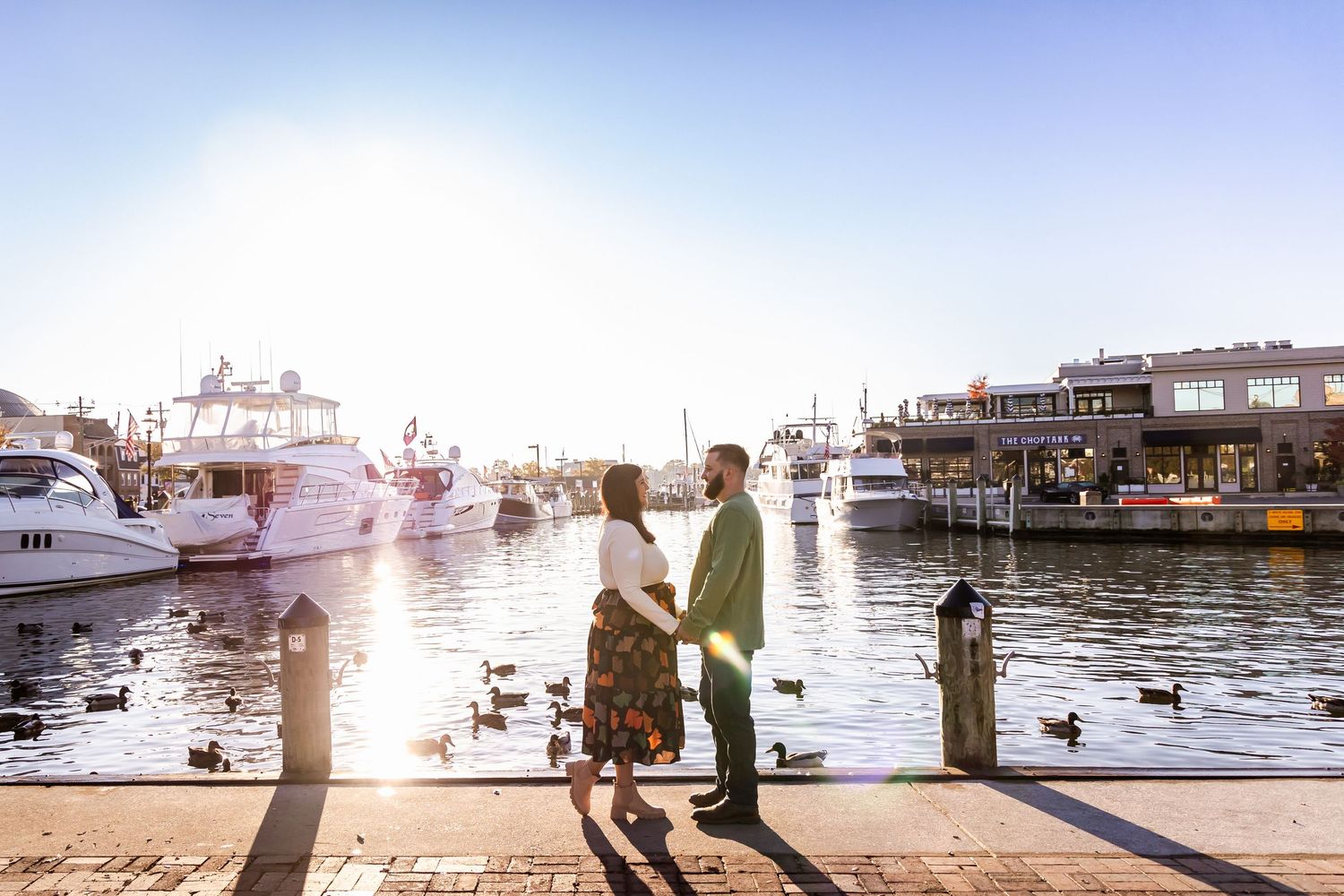 Silhouetted people walking along a waterfront pier at sunset with boats and harbor buildings in the background.