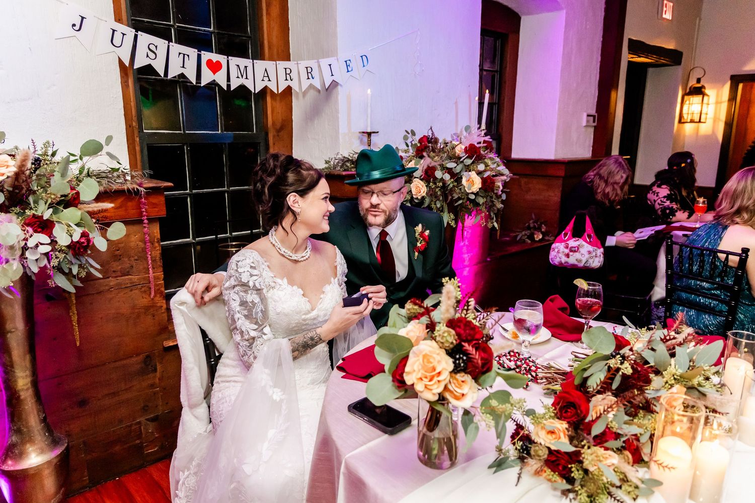 Reception hall decorated with pink lighting and floral centerpieces on white tablecloths.