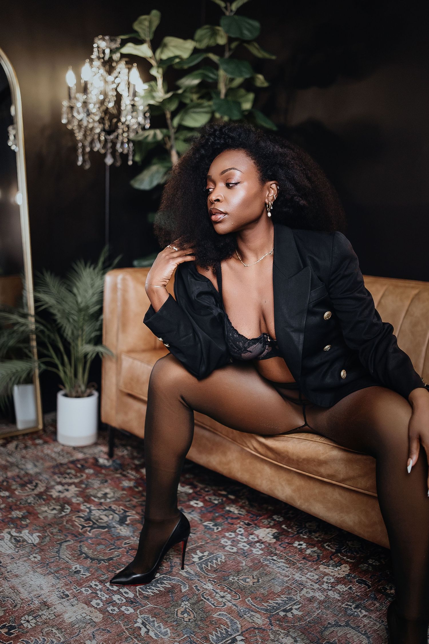Artistic boudoir photoshoot featuring elegant poses on a brown leather couch with chandelier and plants in background.