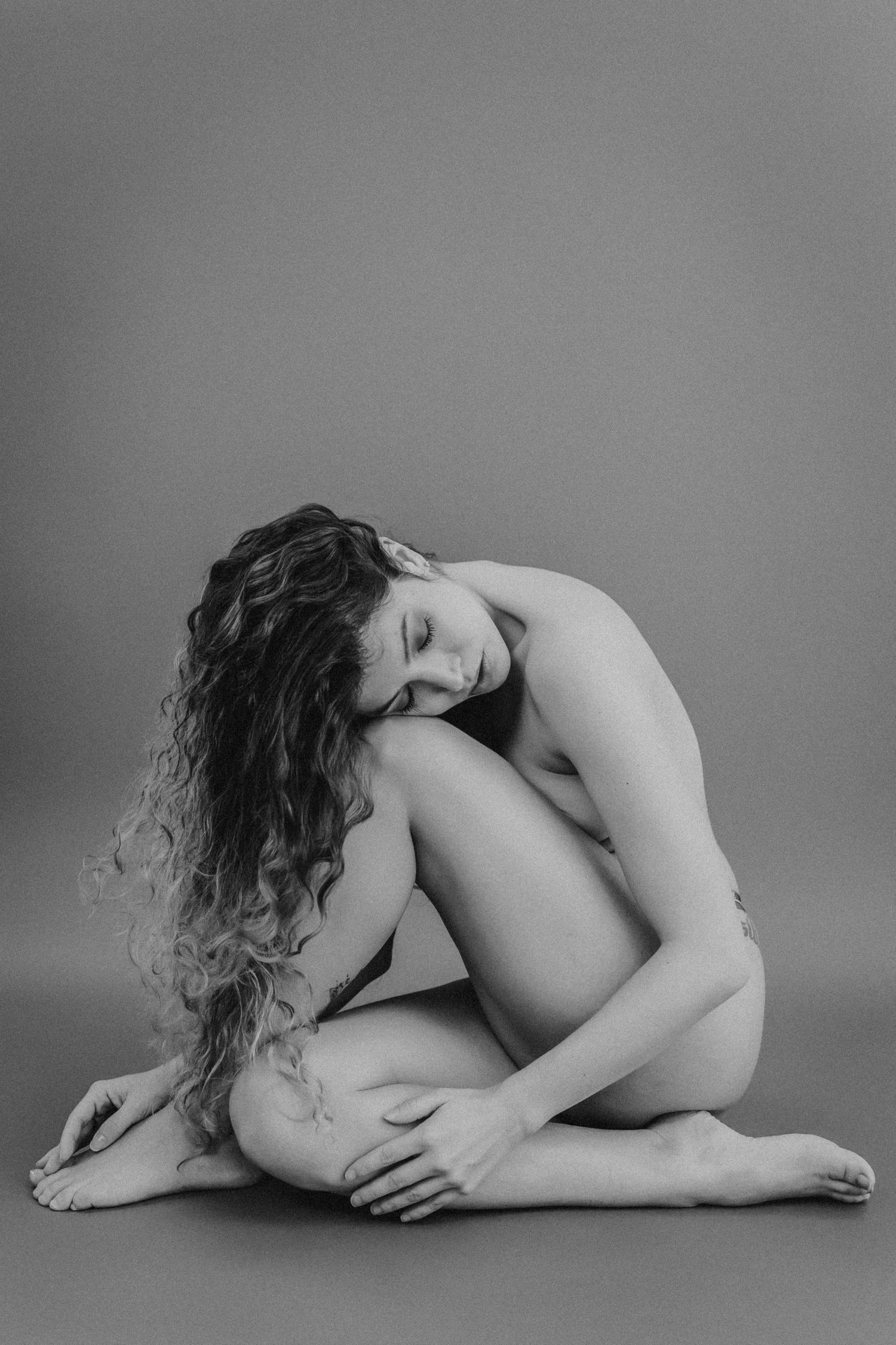 Black and white artistic nude photograph showing a curled up figure with long curly hair against a plain background.