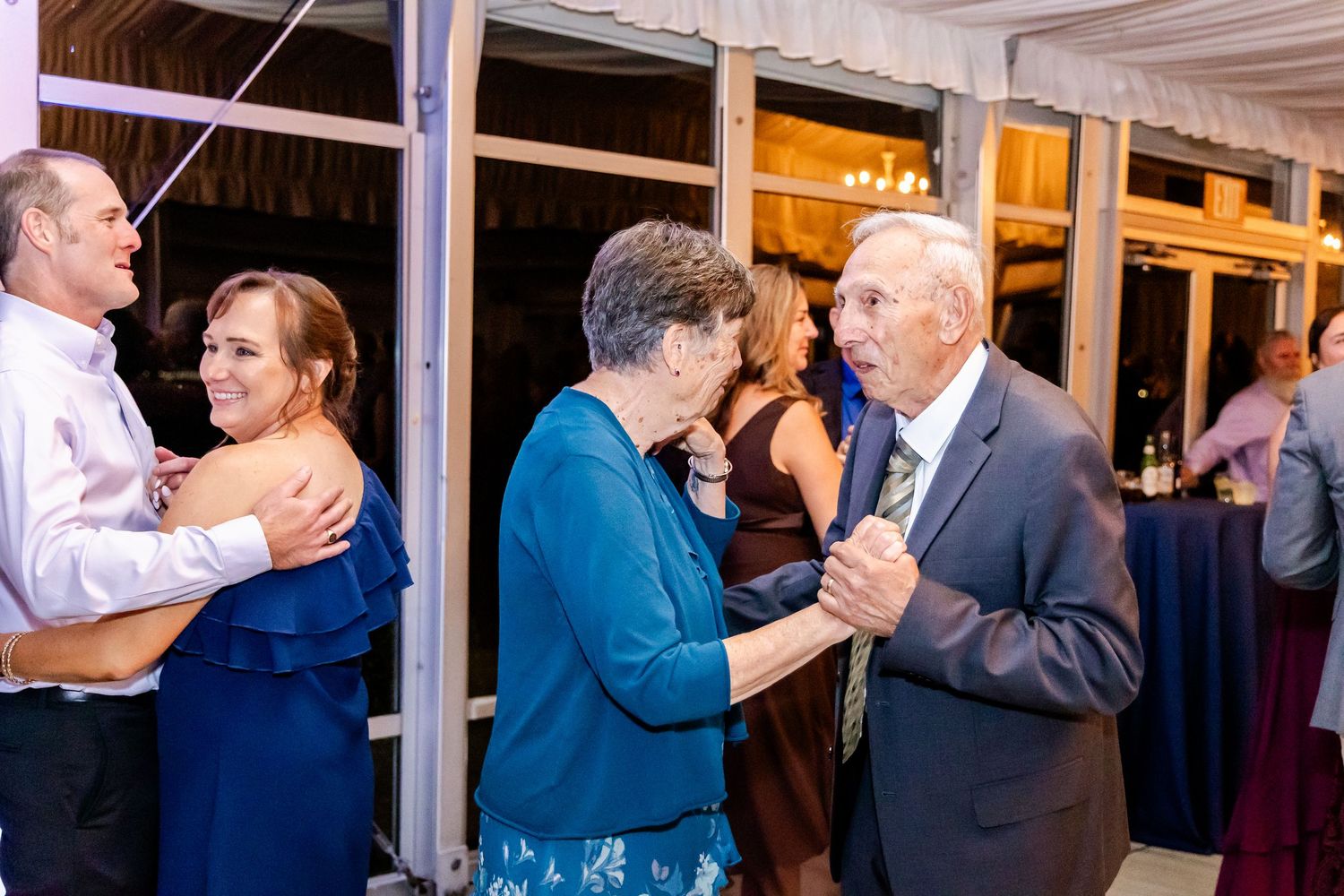 Couples dancing together at an indoor event venue with white ceiling beams and warm lighting.