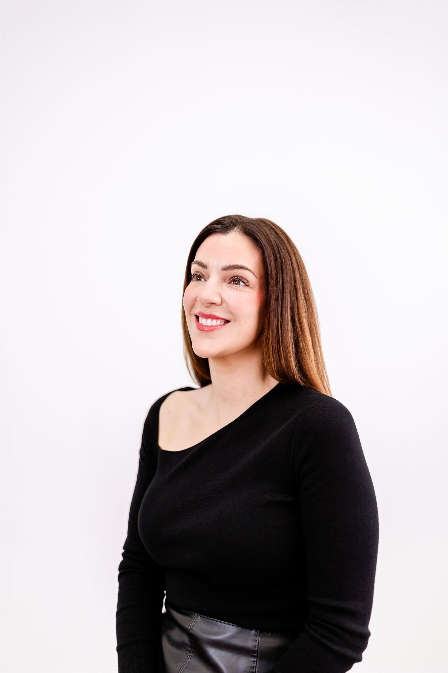 Smiling woman with long brown hair wearing a black long-sleeve top and leather skirt against a white background.
