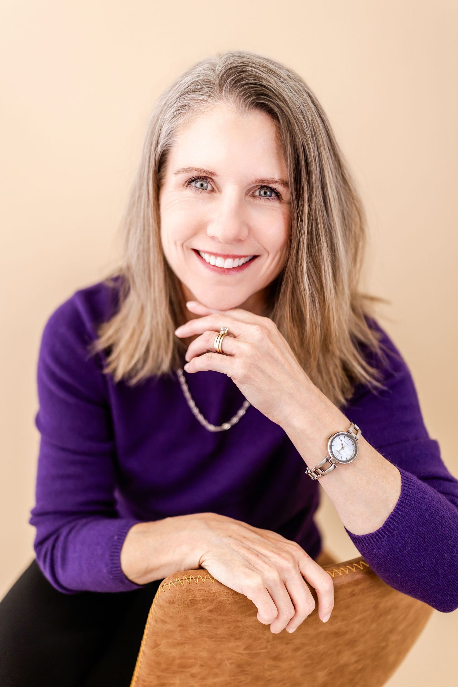 Smiling woman in purple sweater with gray hair, wearing a watch and rings, posed against a beige background.