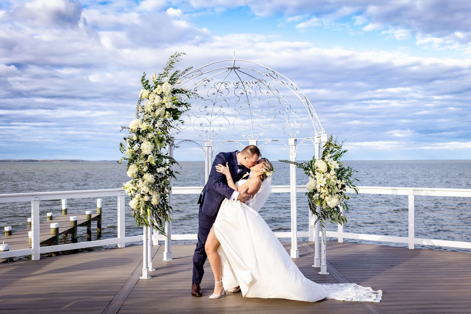 A romantic beach wedding ceremony setup with white floral arrangements and wooden chairs along a pier by the ocean.