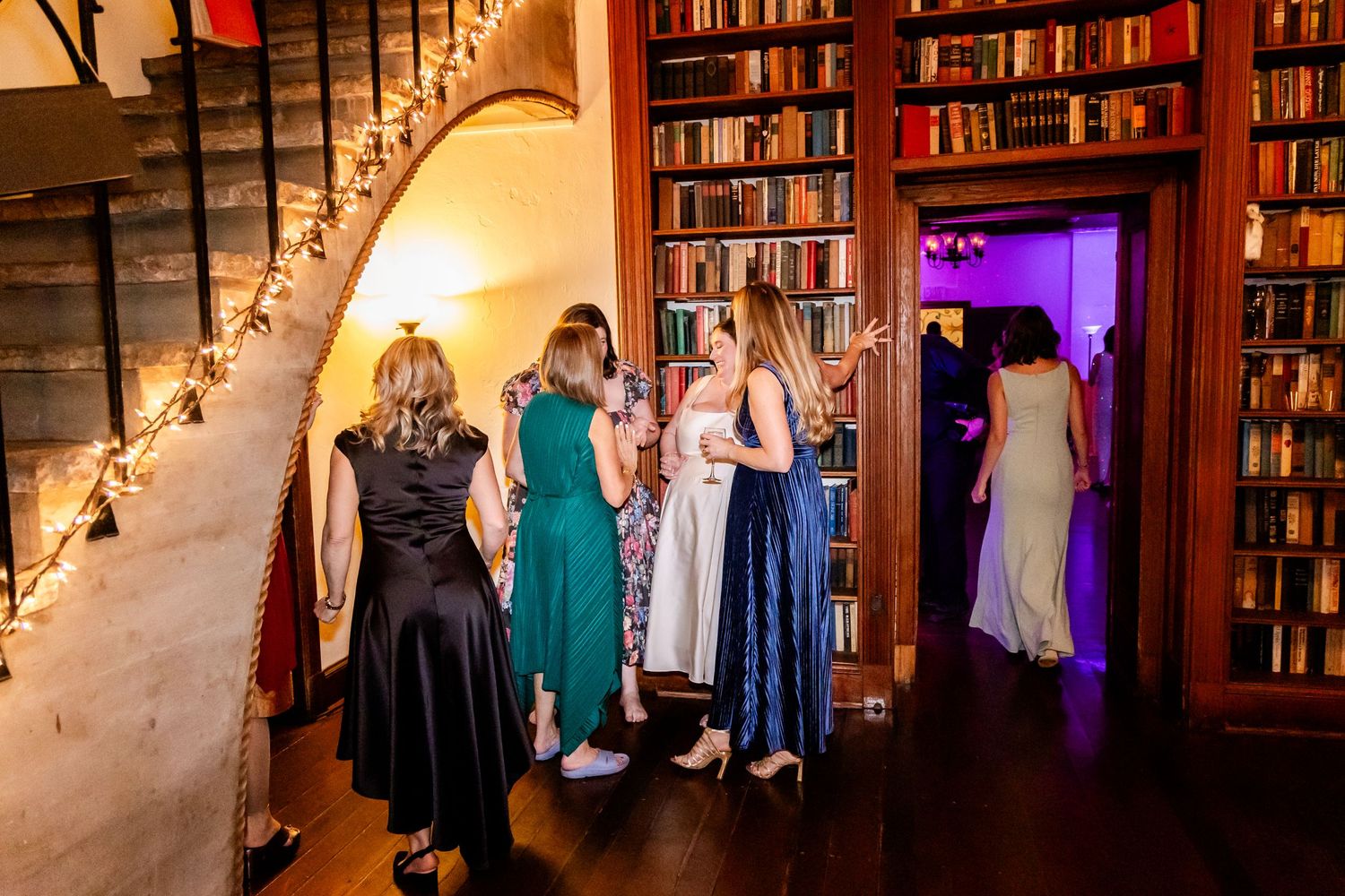 Wedding guests gather in an elegant library space decorated with twinkling lights and floor-to-ceiling bookshelves.