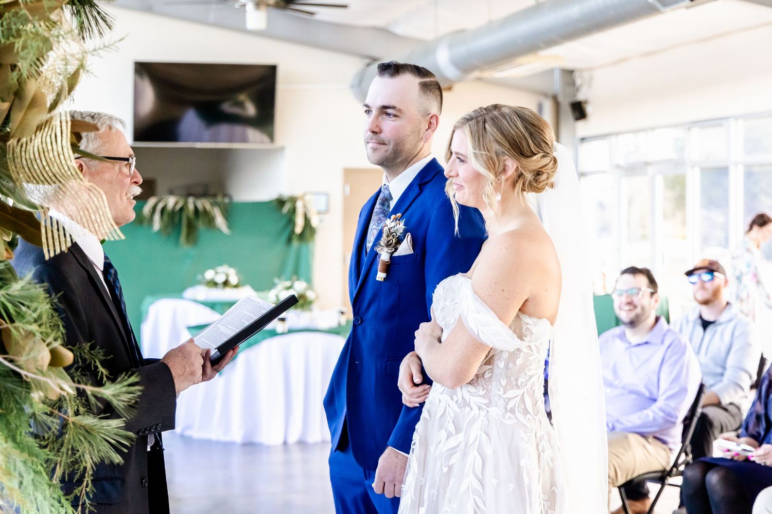 Wedding ceremony taking place in a bright venue with white decor and greenery accents.