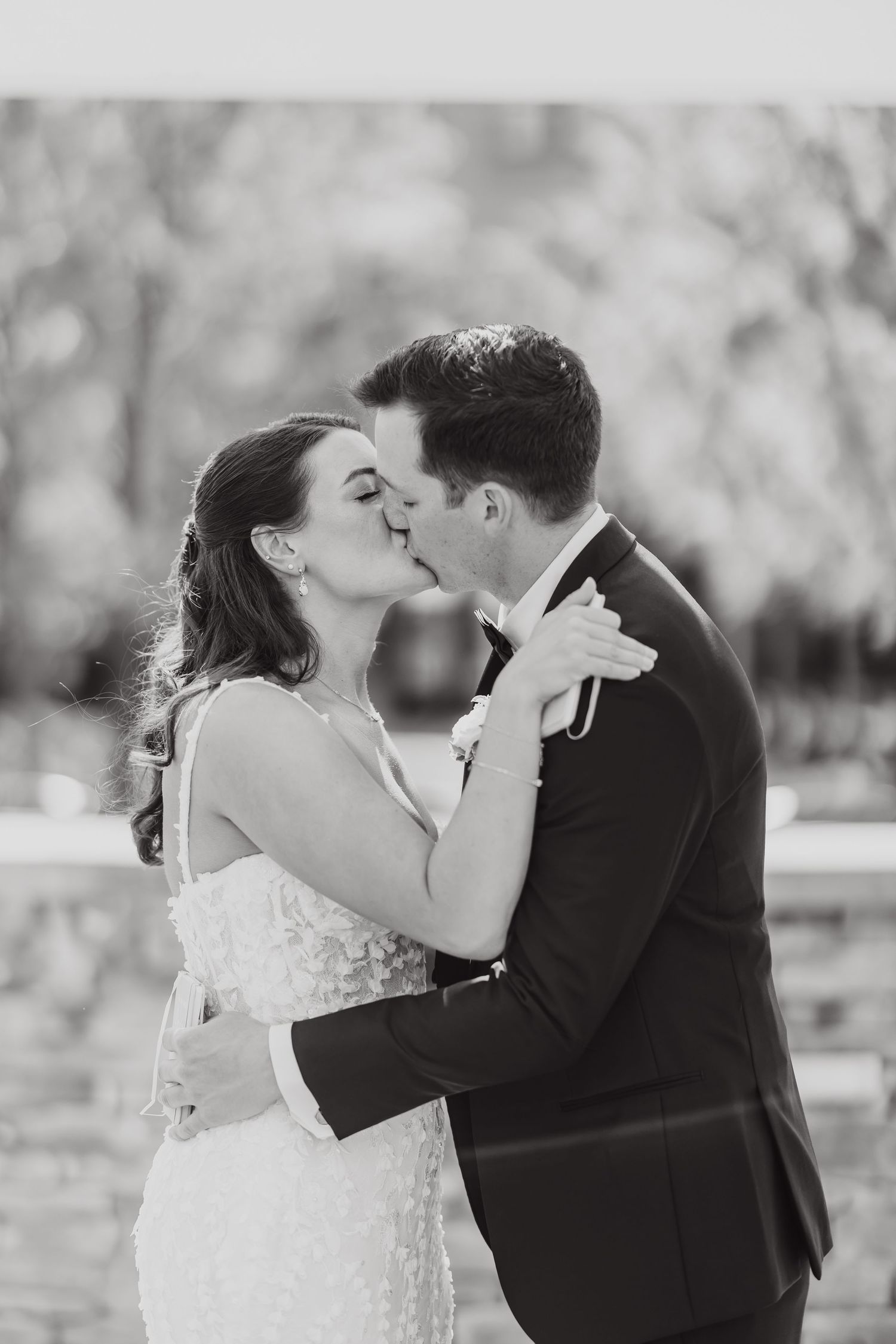 A romantic black and white wedding photo of a couple sharing their first kiss in an outdoor setting.