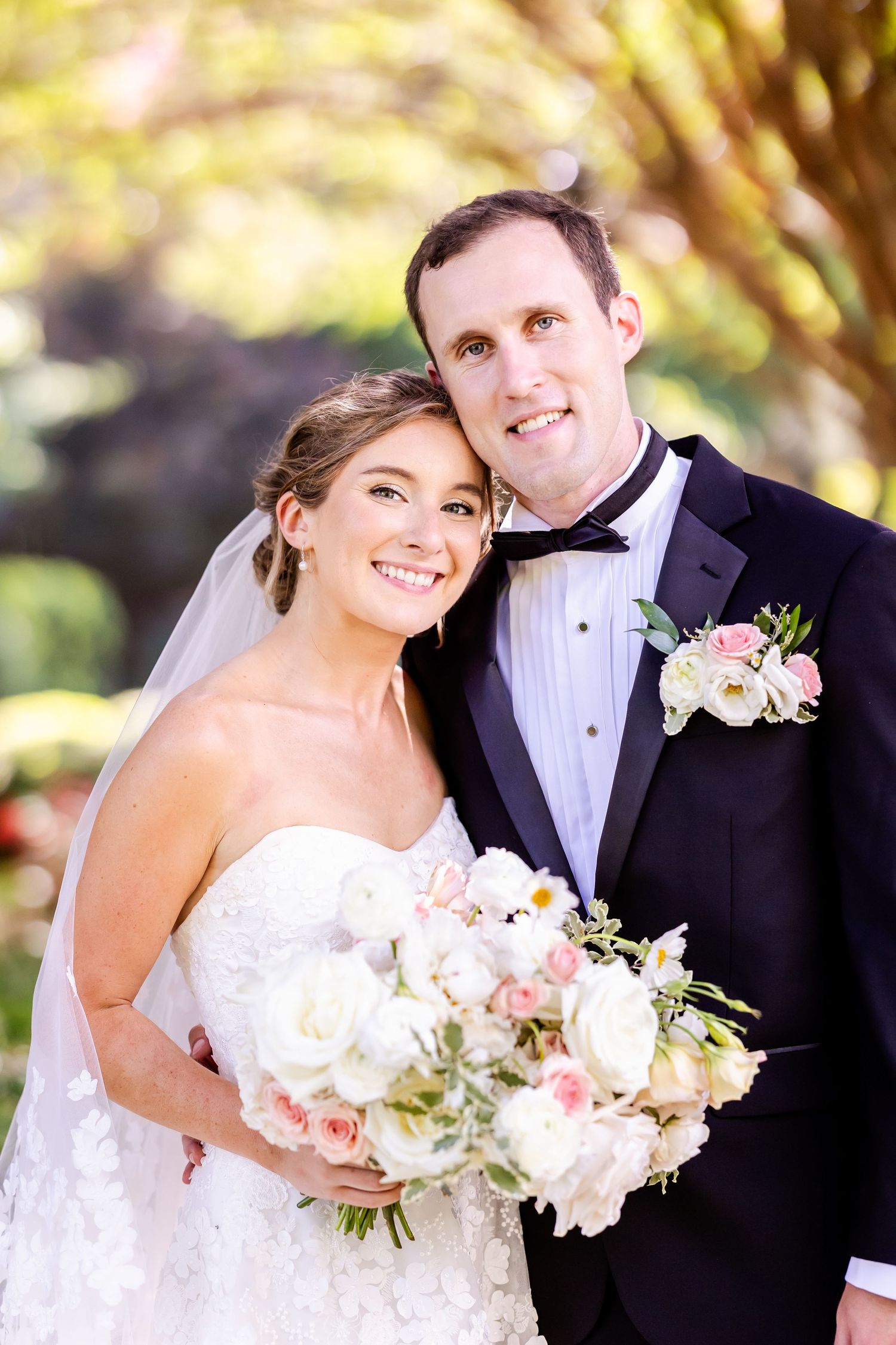 Happy bride in white gown holding pink bouquet poses with groom under sunlit trees.